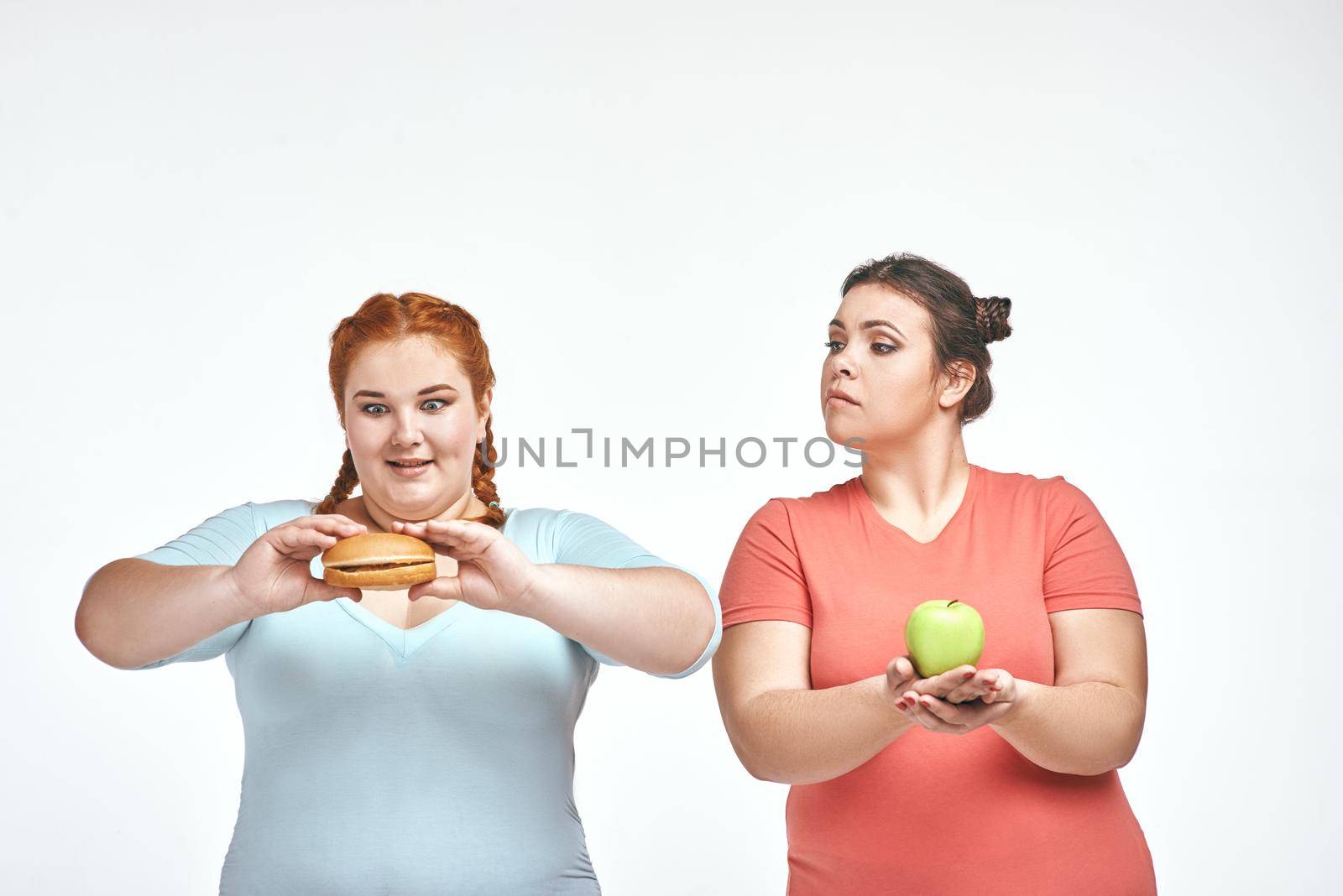 Funny picture of amusing chubby women on white background. One woman holding a sandwich, the other holding an apple.