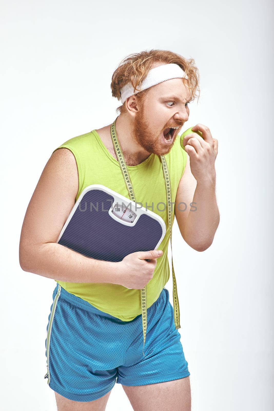 Funny picture of red haired, bearded, plump man on white background. Man holding an apple and scales