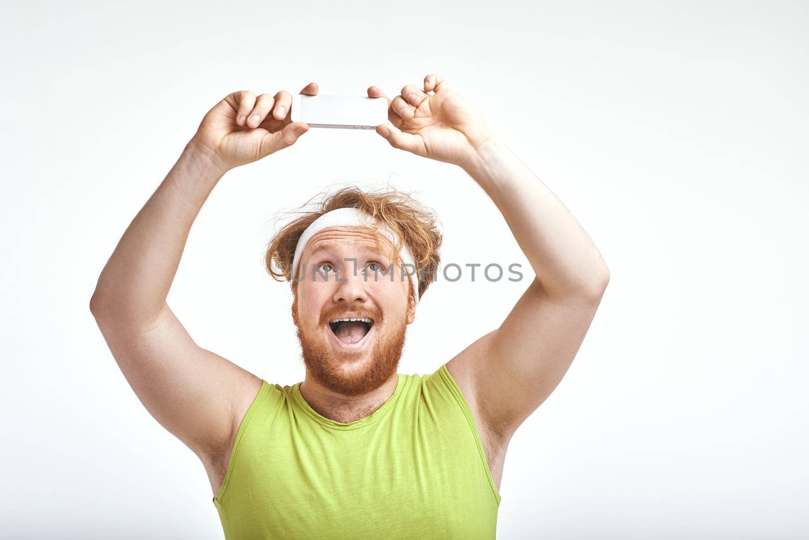 Funny picture of red haired, bearded, plump man on white background. Man wearing sportswear. Man smiling and taking a selfie