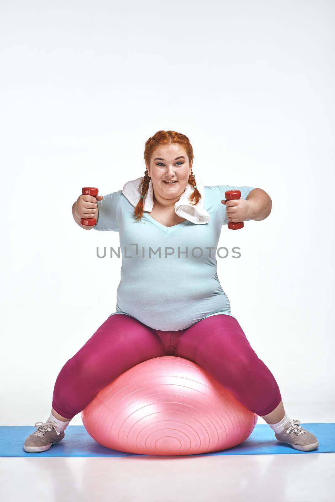 Funny picture of amusing, red haired, chubby woman on white background. Woman sitting on the ball and keep the dumbbells