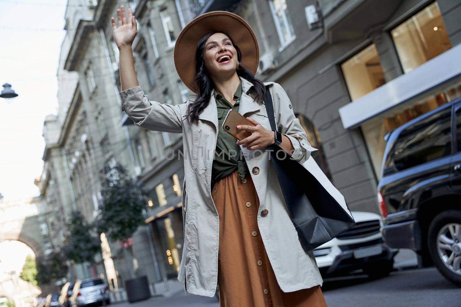Shopping addiction. Young beautiful stylish woman in hat with shopping bag holding her smartphone and waving to friend while standing on the city street by friendsstock