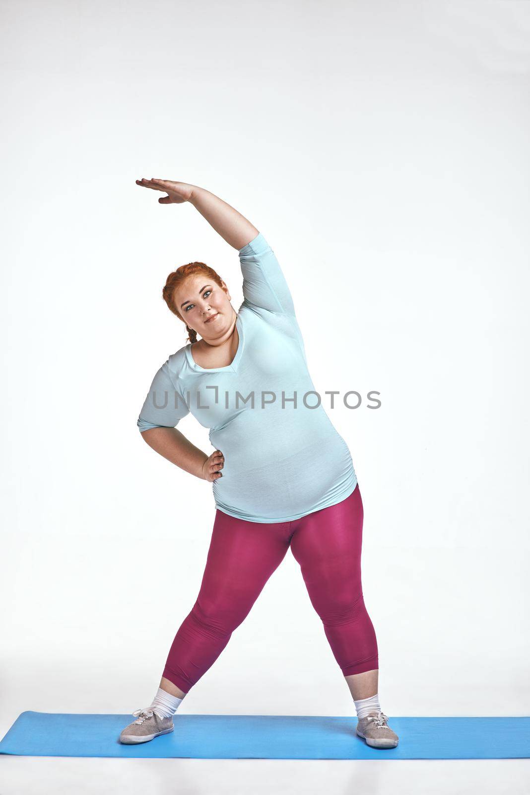Funny picture of amusing, red haired, chubby woman on white background. Woman trains on the mat.