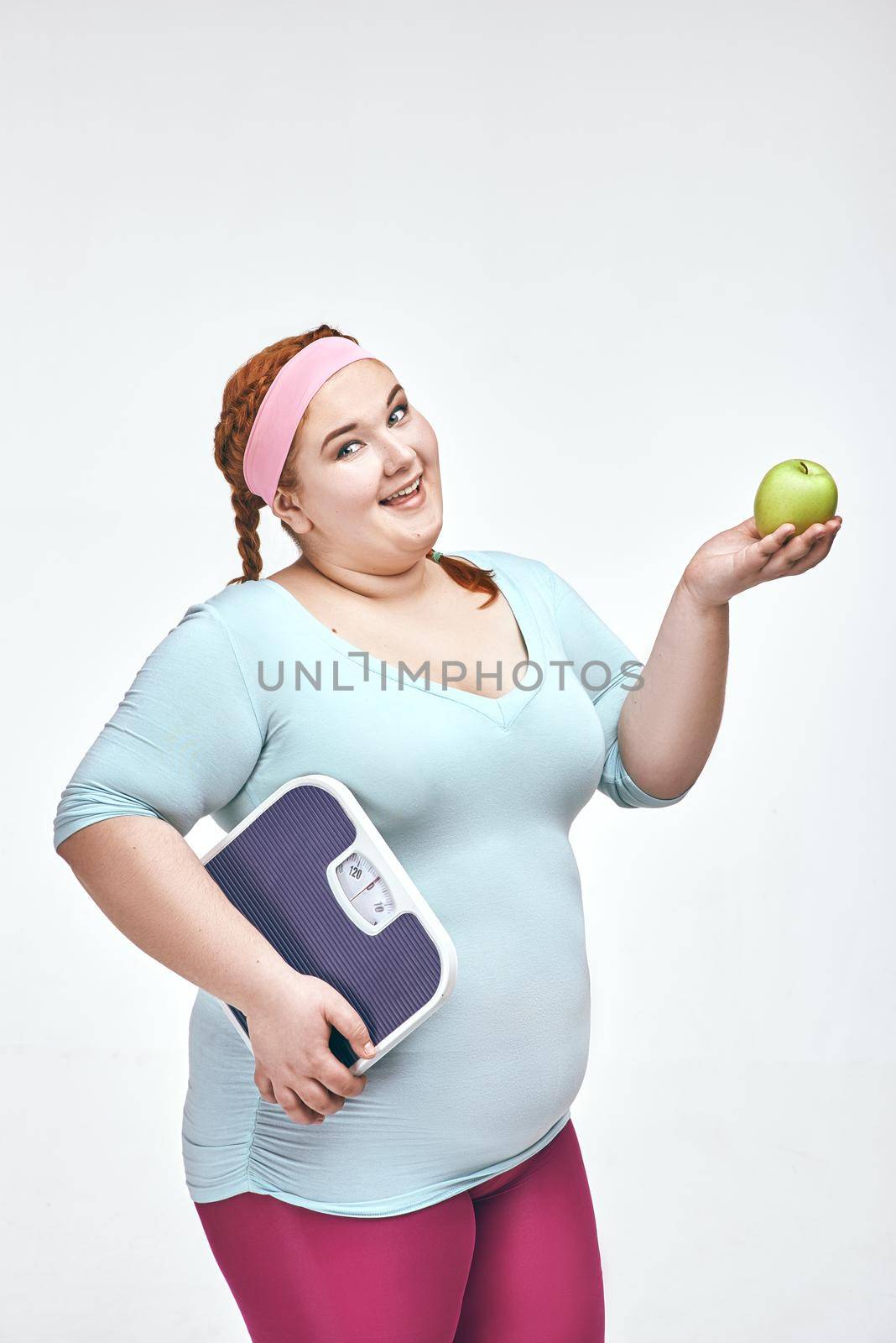 Funny picture of amusing, red haired, chubby woman on white background. Woman holding an apple and scales.