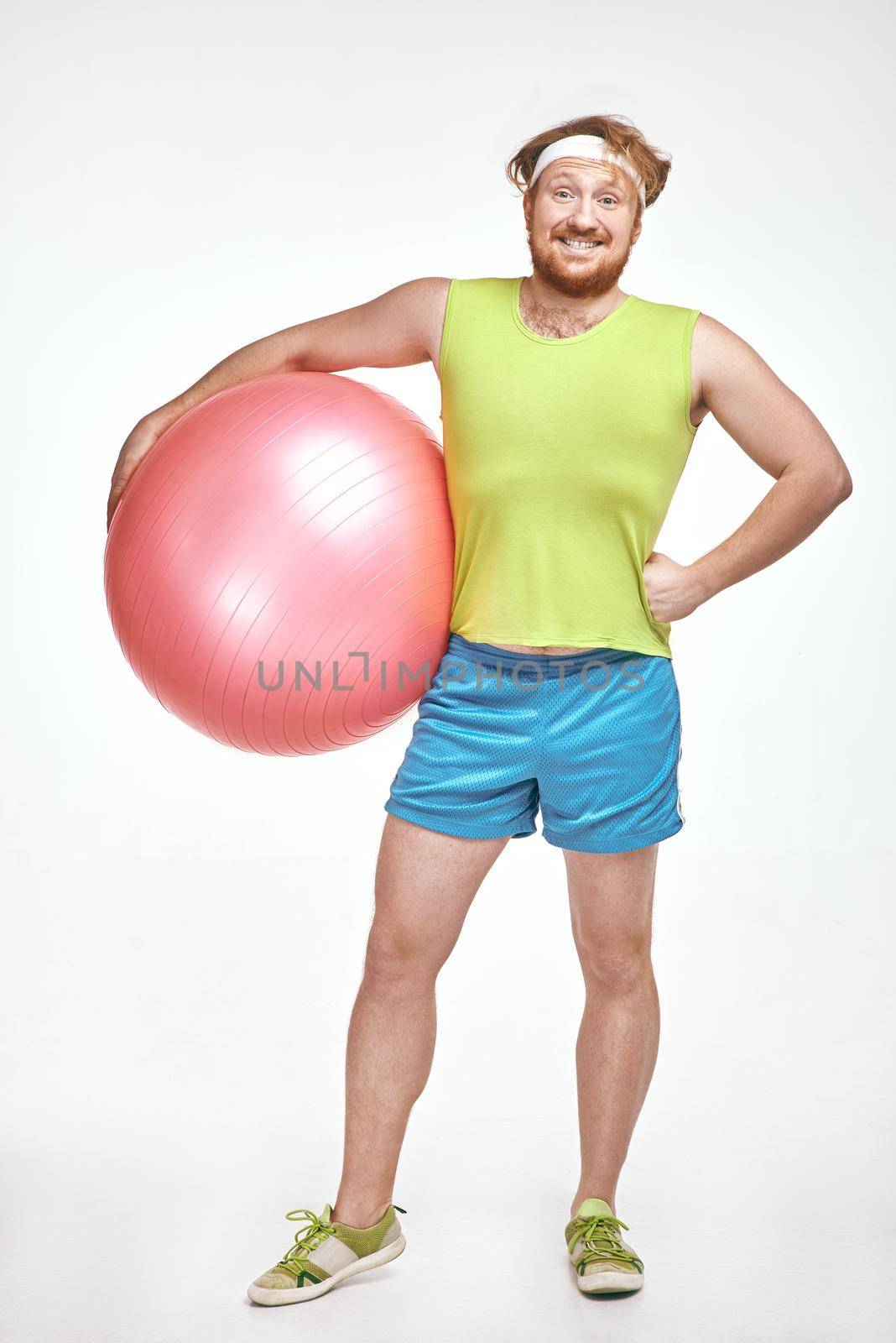 Funny picture of red haired, bearded, plump man on white background. Man wearing sportswear. Man holding fitness ball