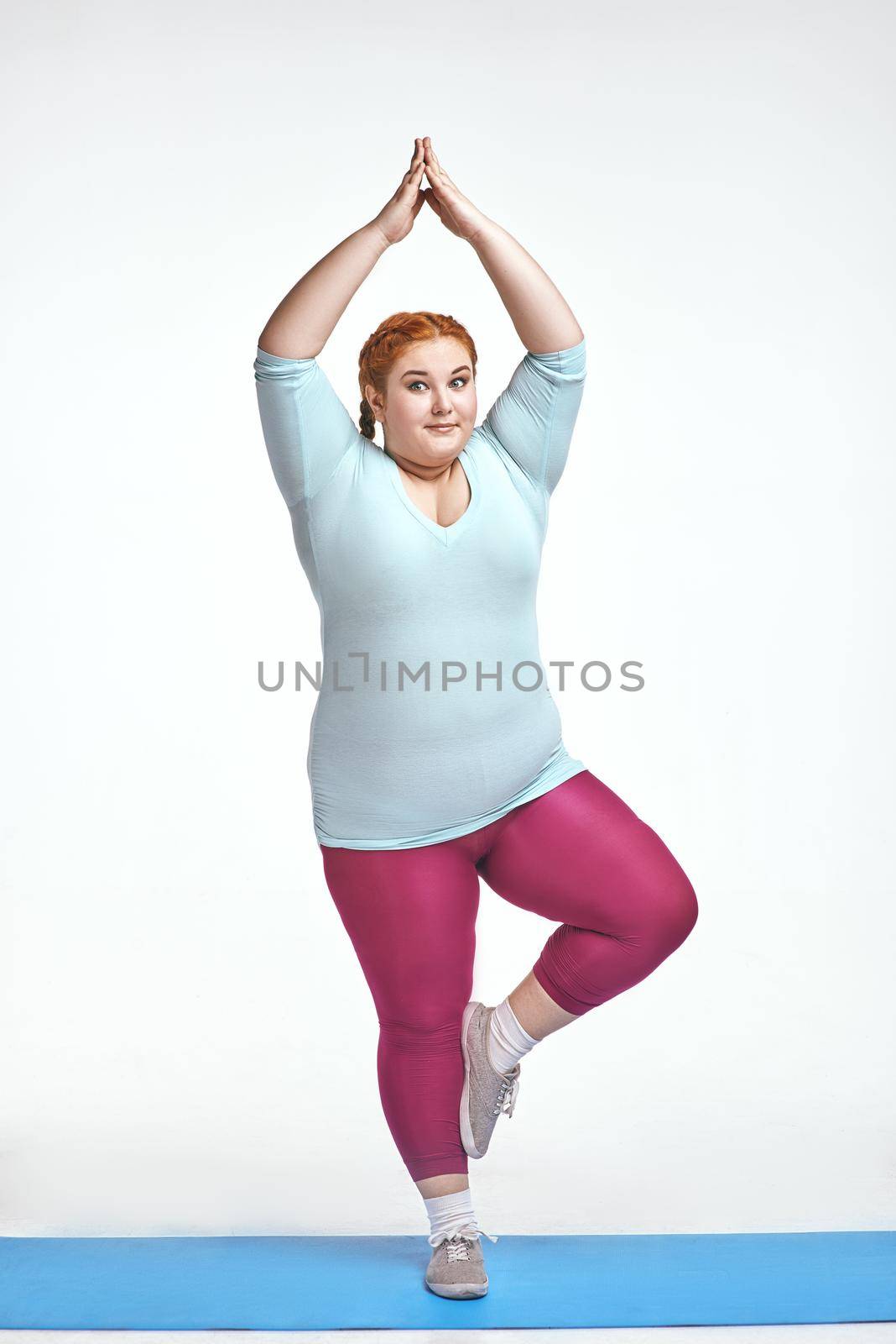 Funny picture of amusing, red haired, chubby woman on white background. Woman trains on the mat.