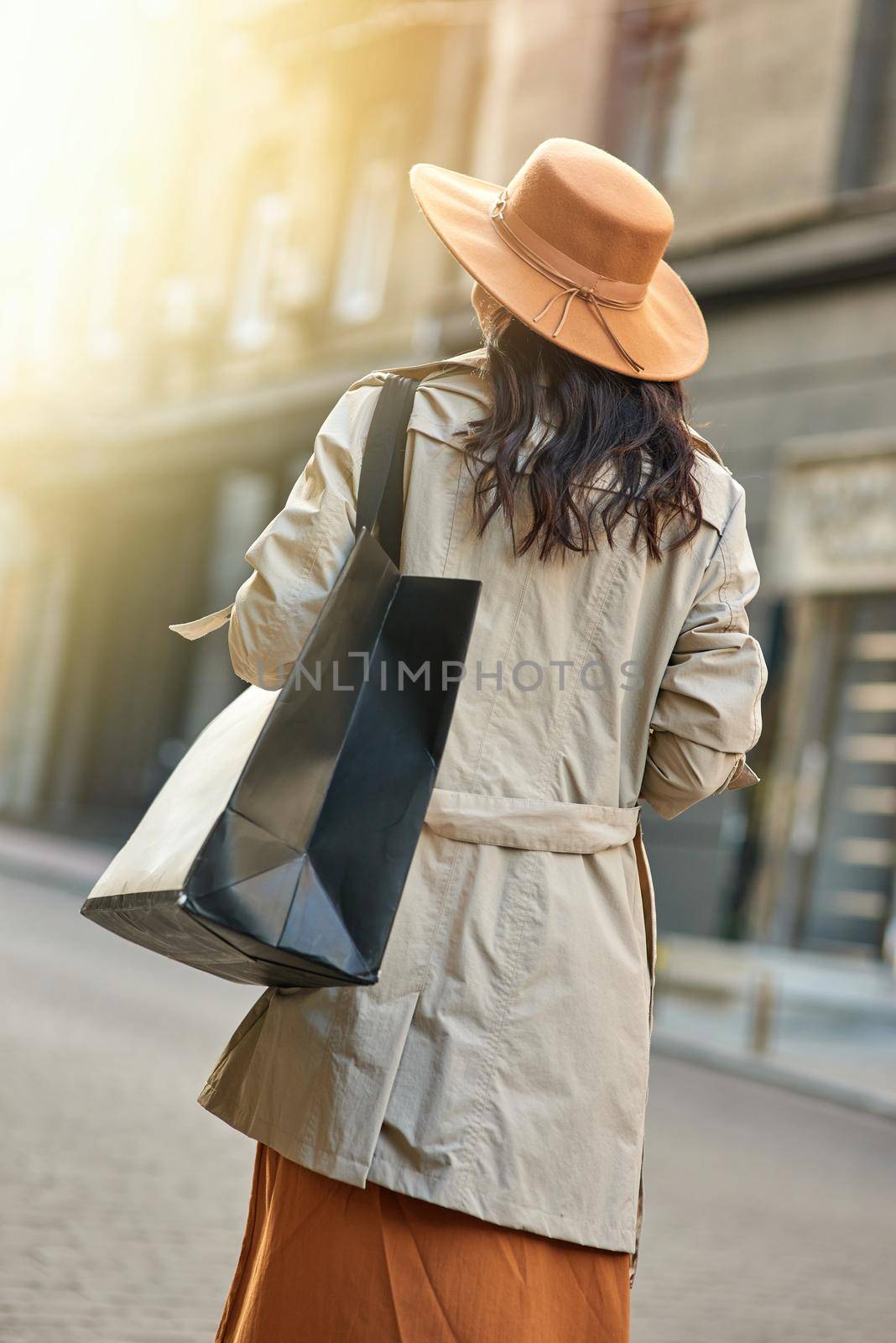 Rear view of a stylish woman wearing autumn grey coat and hat with big shopping bag walking city streets. Shopaholism, fashion, people lifestyle concept