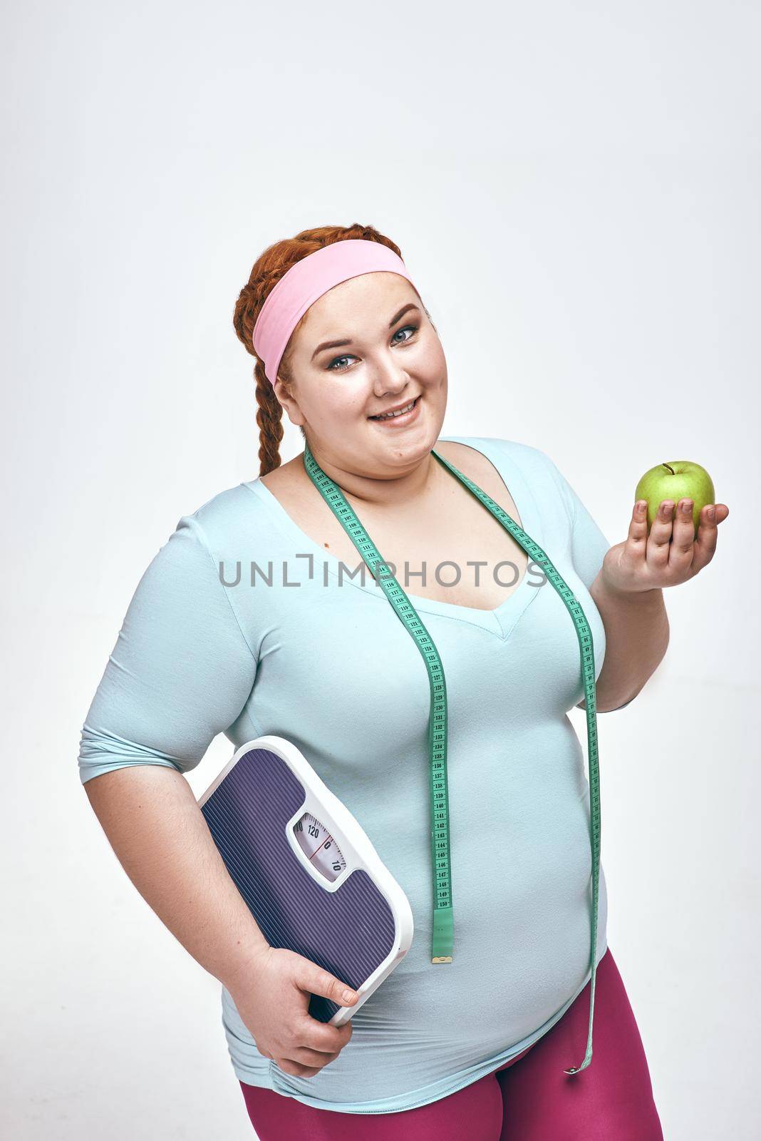 Funny picture of amusing, red haired, chubby woman on white background. Woman holding an apple and scales.