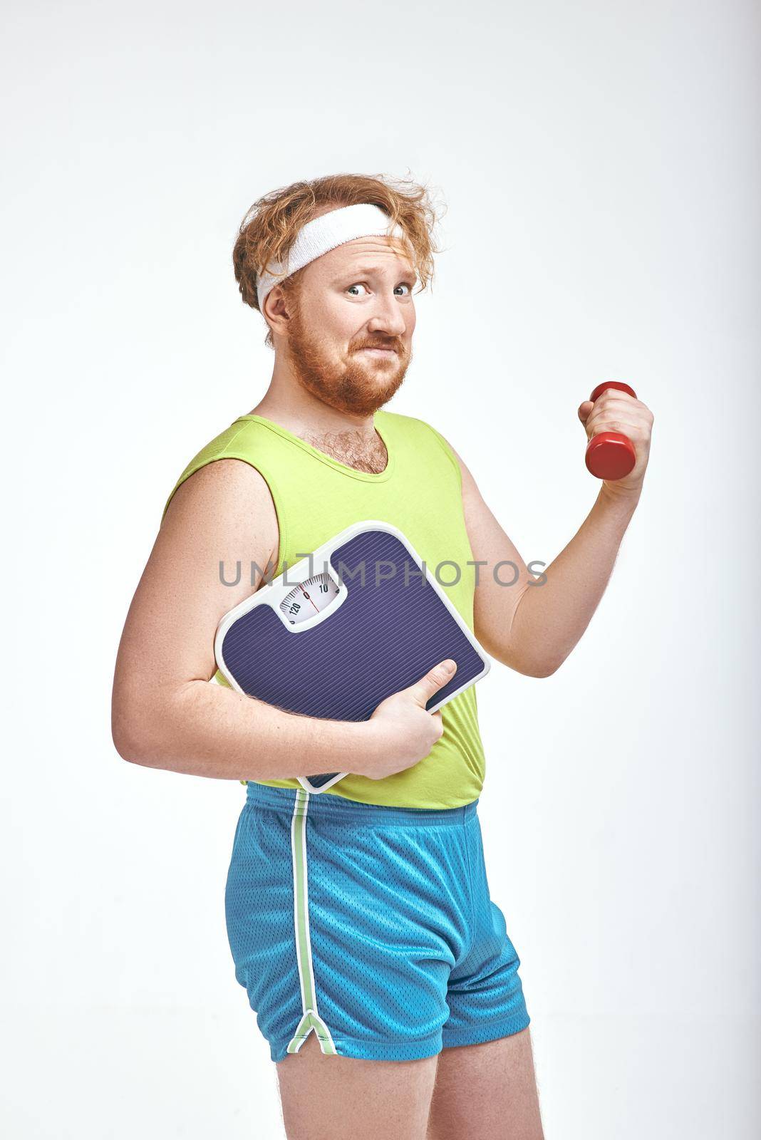Funny picture of red haired, bearded, plump man on white background. Man holding a dumbbell and scales