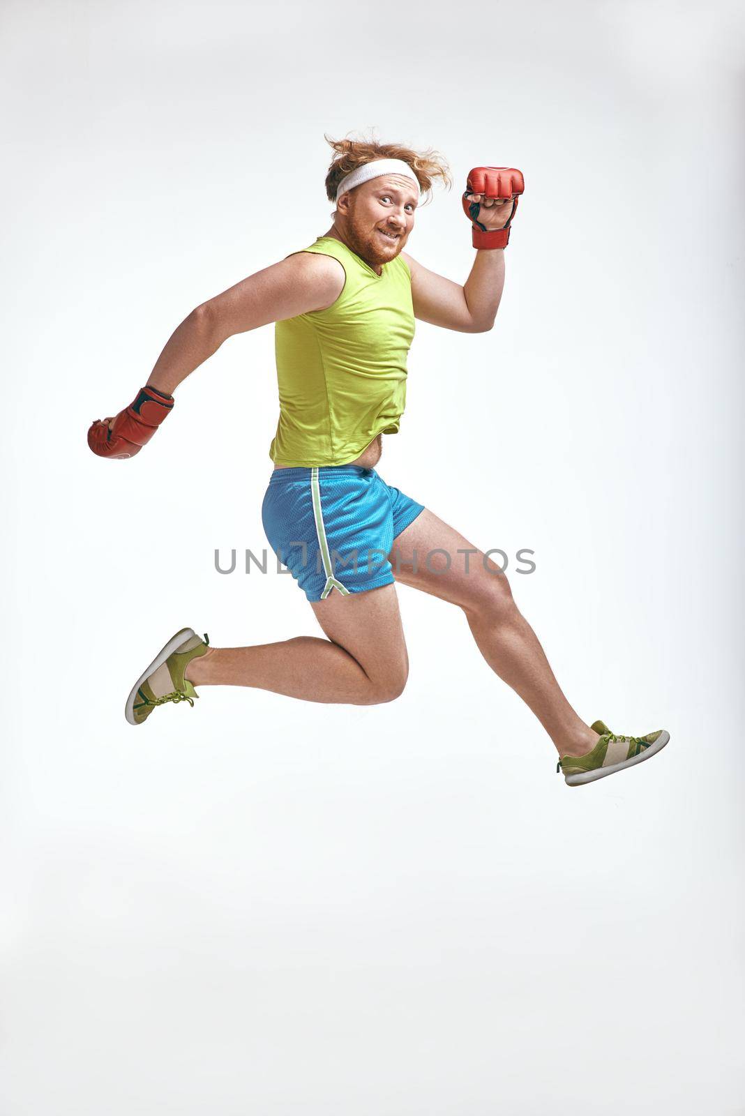Funny picture of red haired, bearded, plump man on white background. Man wearing sportswear and red boxing gloves