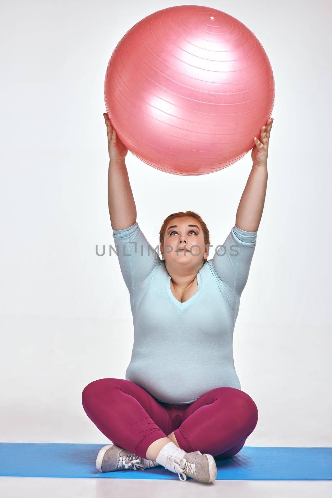Funny picture of amusing, red haired, chubby woman on white background. Woman is sitting and holding a ball