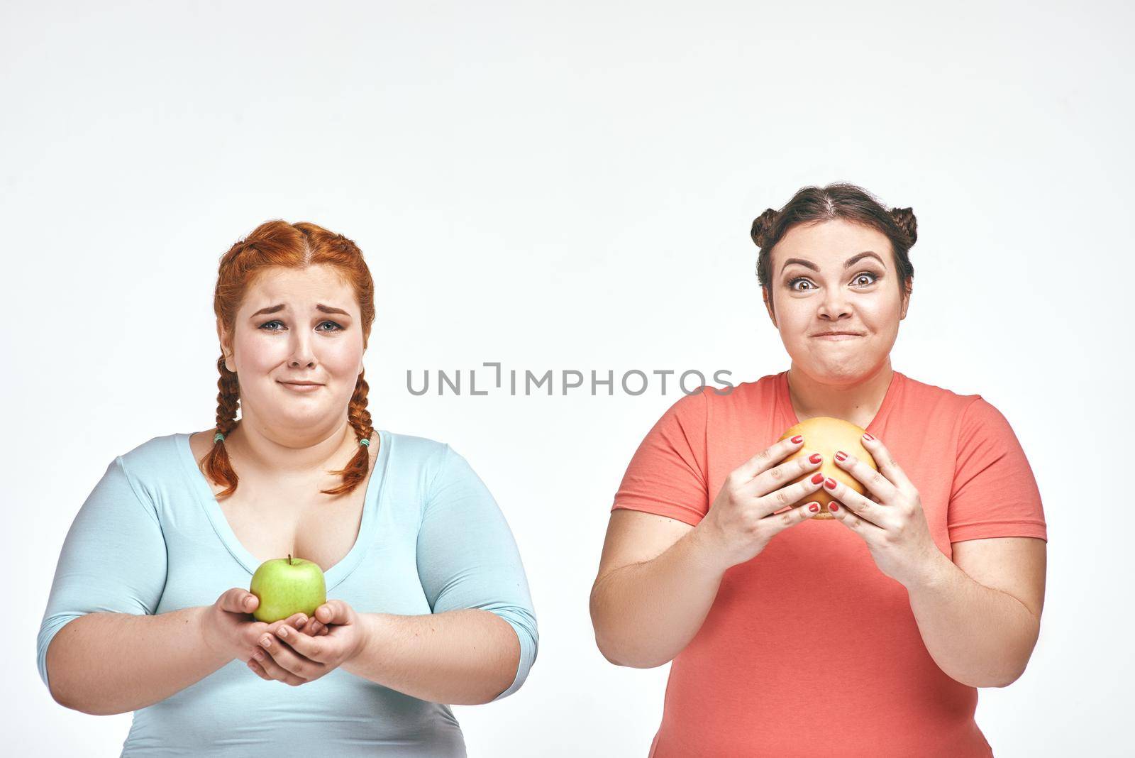 Funny picture of amusing chubby women on white background. One woman holding a sandwich, the other holding an apple.