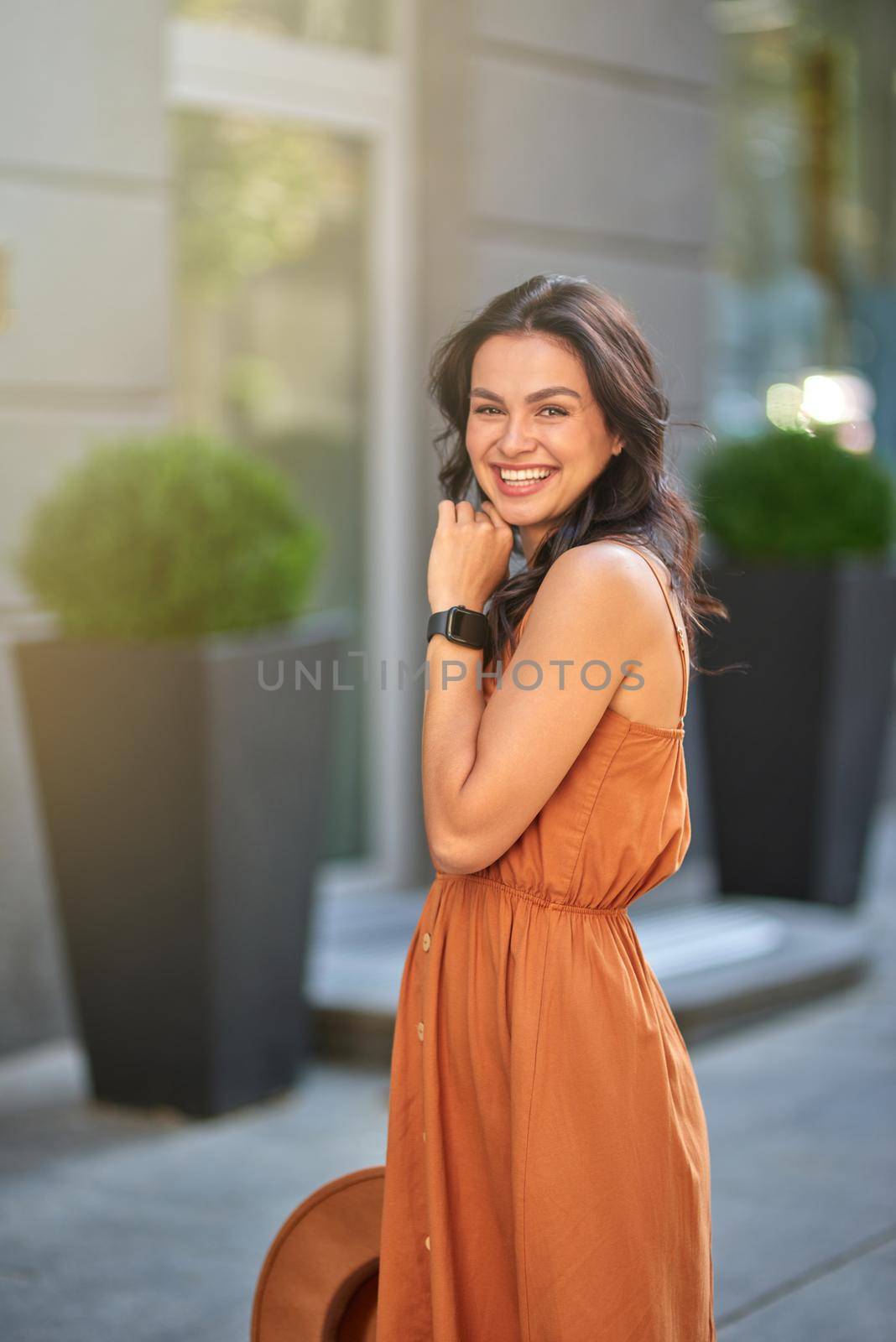 Vertical shot of a young romantic caucasian woman wearing summer dress holding her hat, looking at camera and smiling while walking city streets on a beautiful sunny day by friendsstock