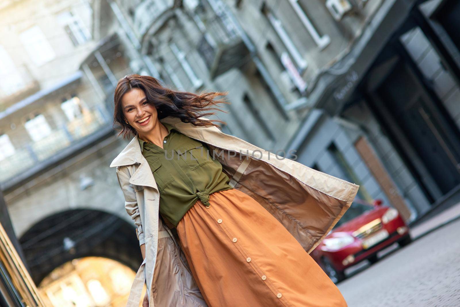 Young excited stylish caucasian woman wearing grey coat looking at camera and smiling while standing on the city street, walking outdoors on a warm autumn day. Beautiful people, fashion concept