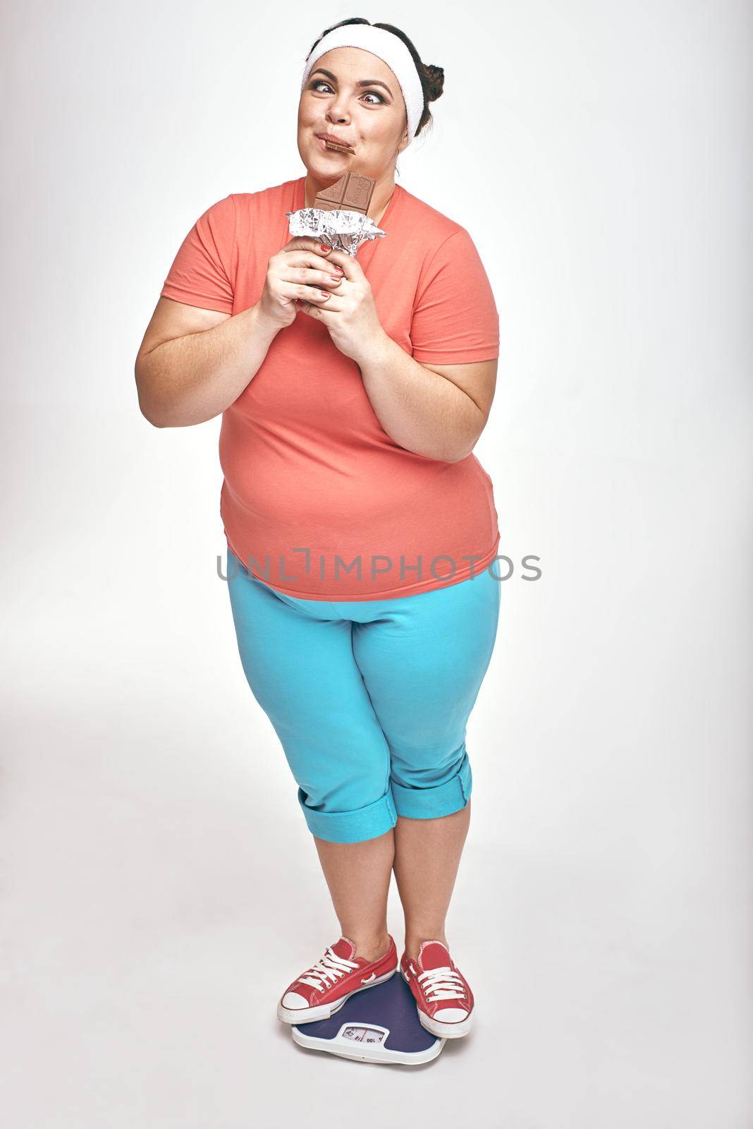 Funny picture of amusing, chubby woman on white background. She is eating chocolate while standing on the scales