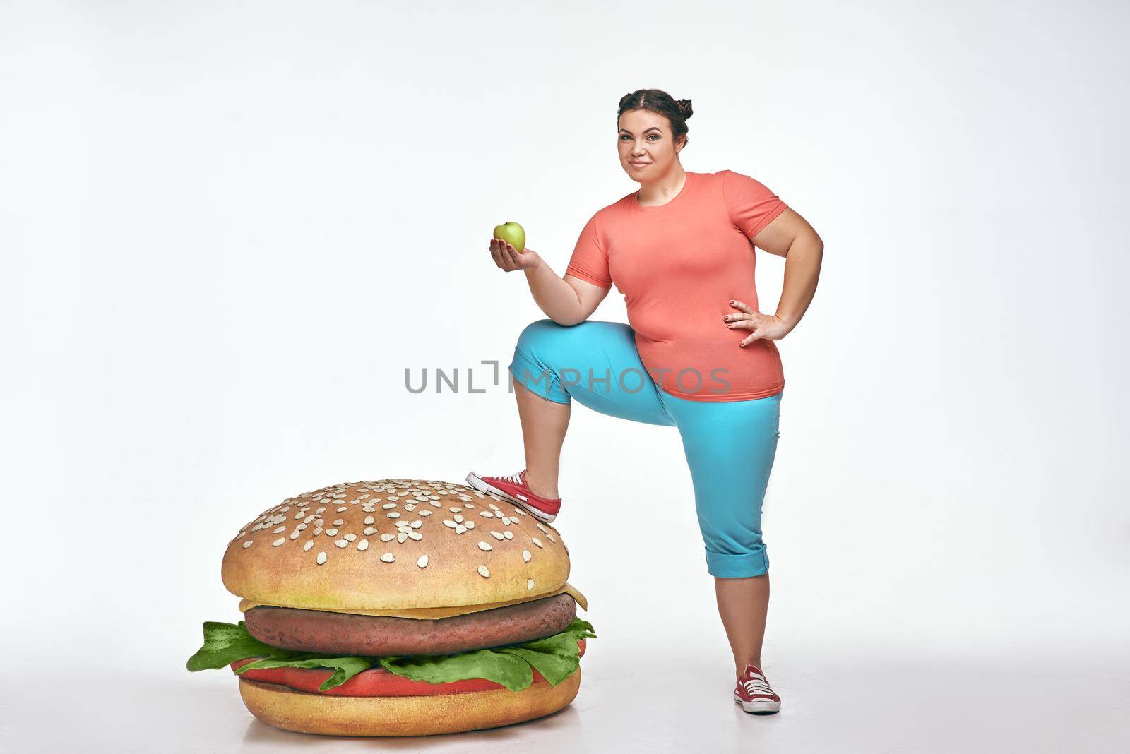 Funny picture of brunette, bearded, plump woman on white background. Woman wearing sportswear. She put her leg to a huge sandwich