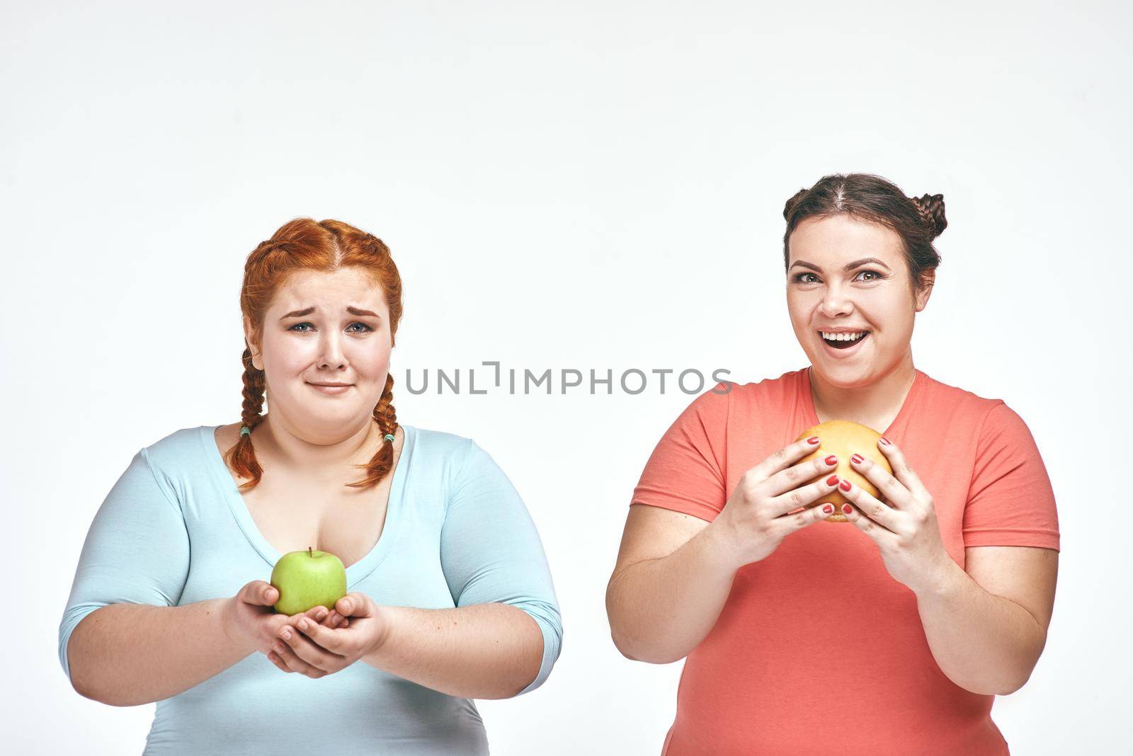 Funny picture of amusing chubby women on white background. One woman holding a sandwich, the other holding an apple.