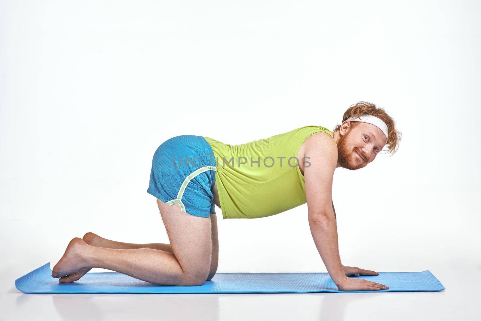 Funny picture of amusing, red haired, chubby man on white background. Man trains on the mat.