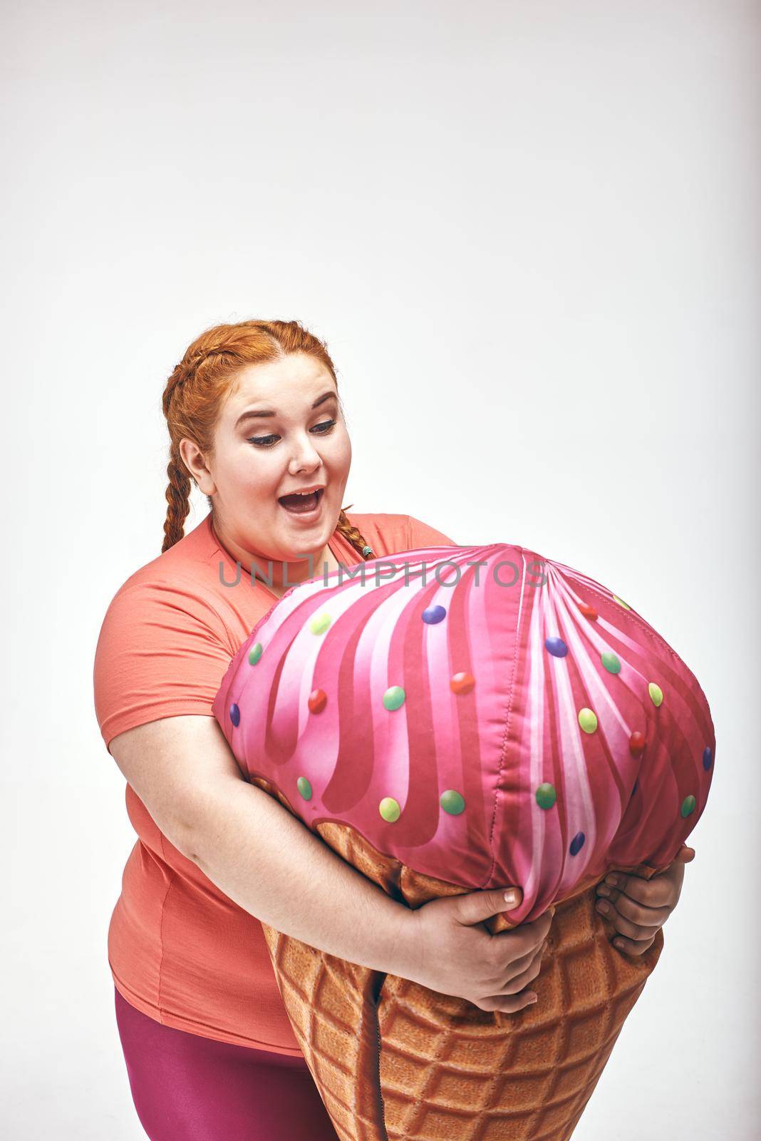 Funny picture of amusing, red haired, chubby woman on white background. Woman is holding a huge ice cream