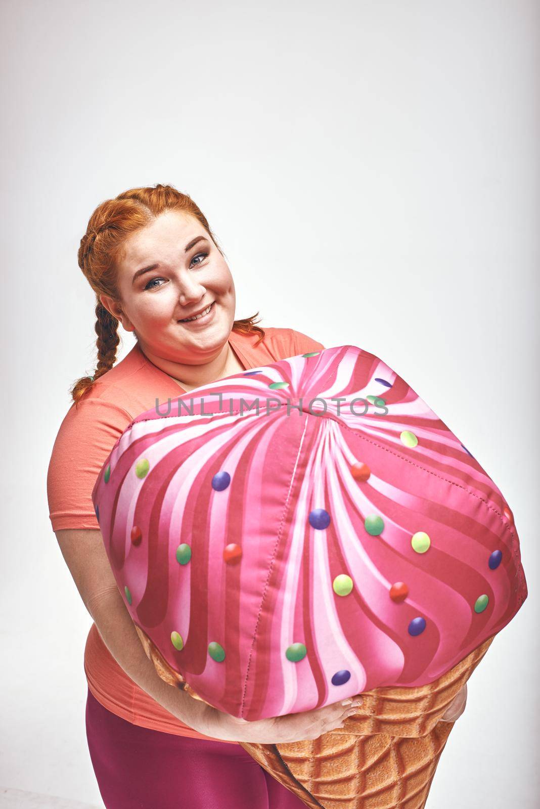 Close-up funny picture of amusing, red haired, chubby woman on white background. Woman is holding a huge ice cream