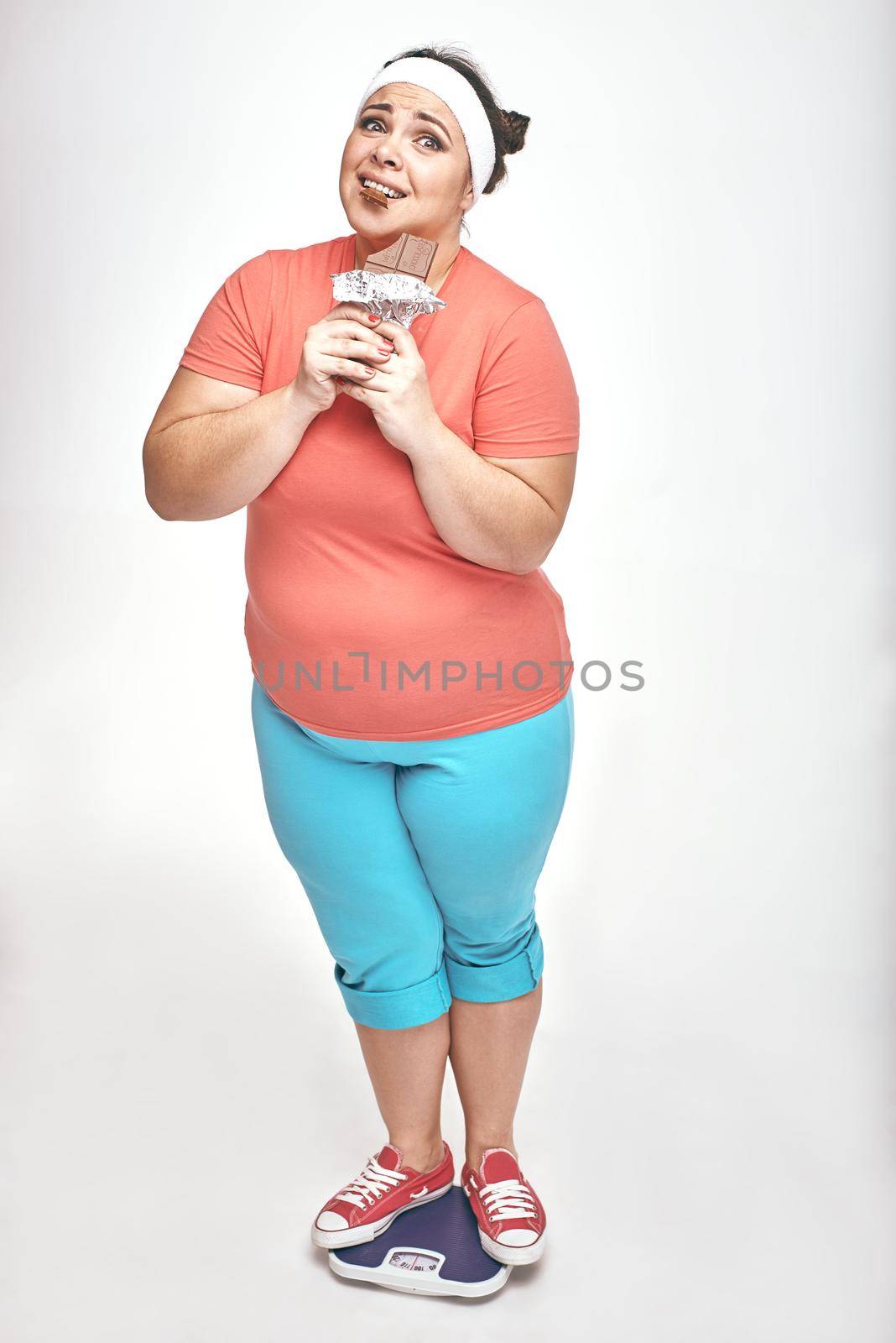 Funny picture of amusing, chubby woman on white background. She is eating chocolate while standing on the scales