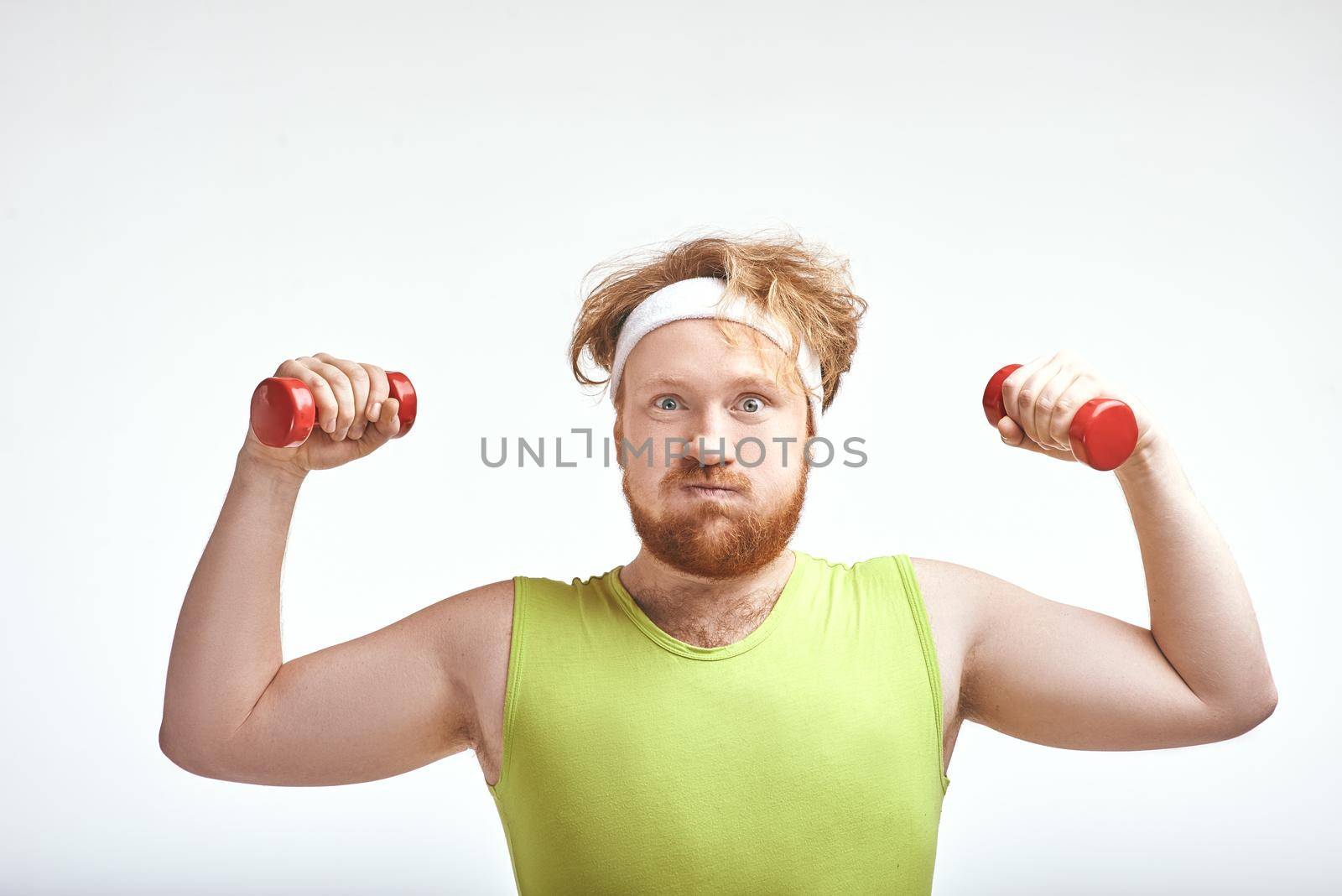 Funny picture of red haired, bearded, plump man on white background. Man wearing sportswear. Man holding the dumbbells