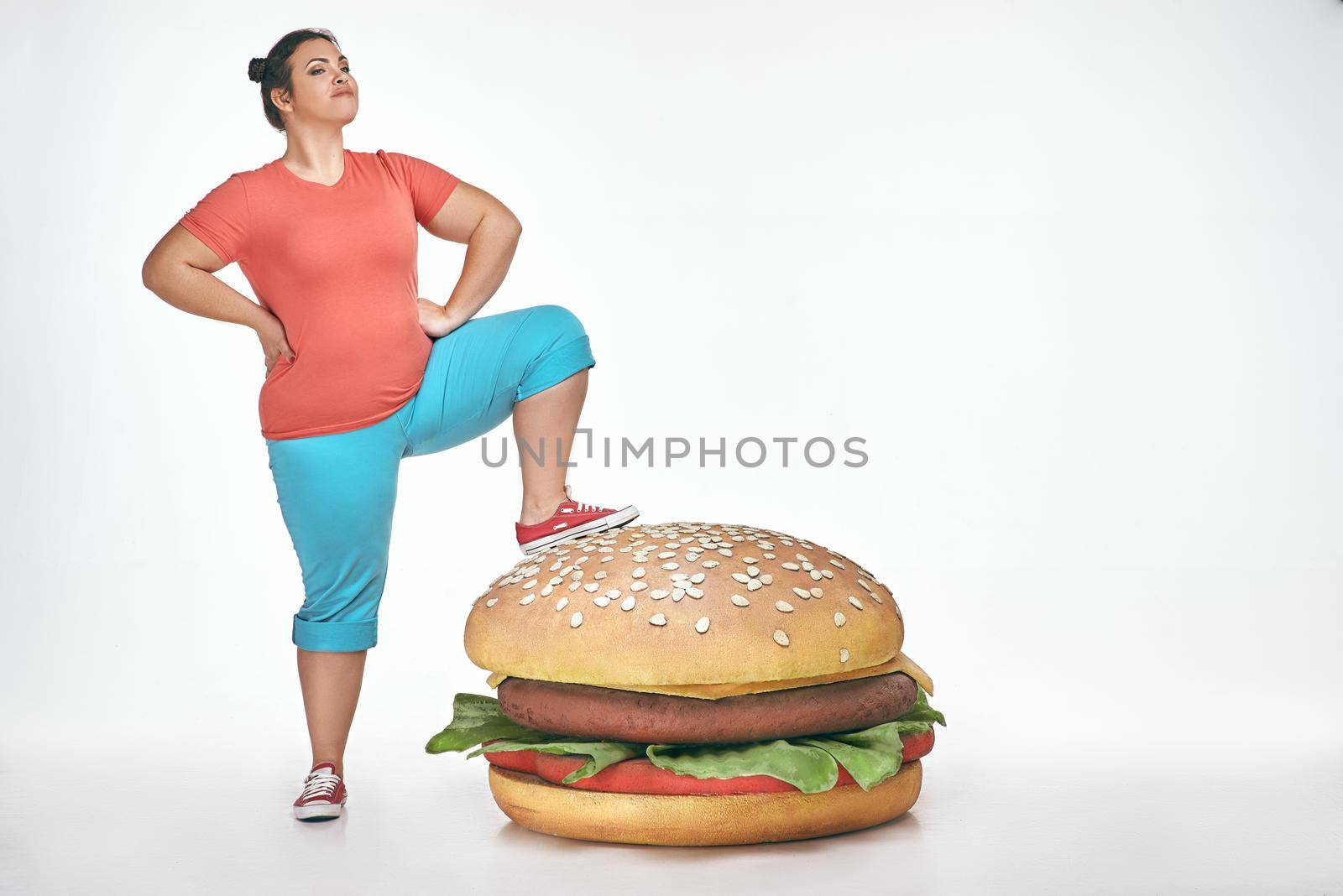 Funny picture of brunette, bearded, plump woman on white background. Woman wearing sportswear. She put her leg to a huge sandwich