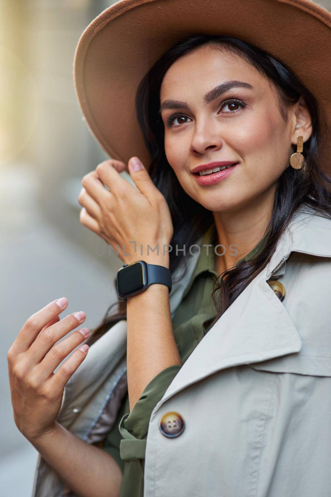 Portrait of attractive stylish woman wearing grey coat and hat looking aside while standing on the city street, enjoying walking on warm autumn day. Fashion and beautiful people concept