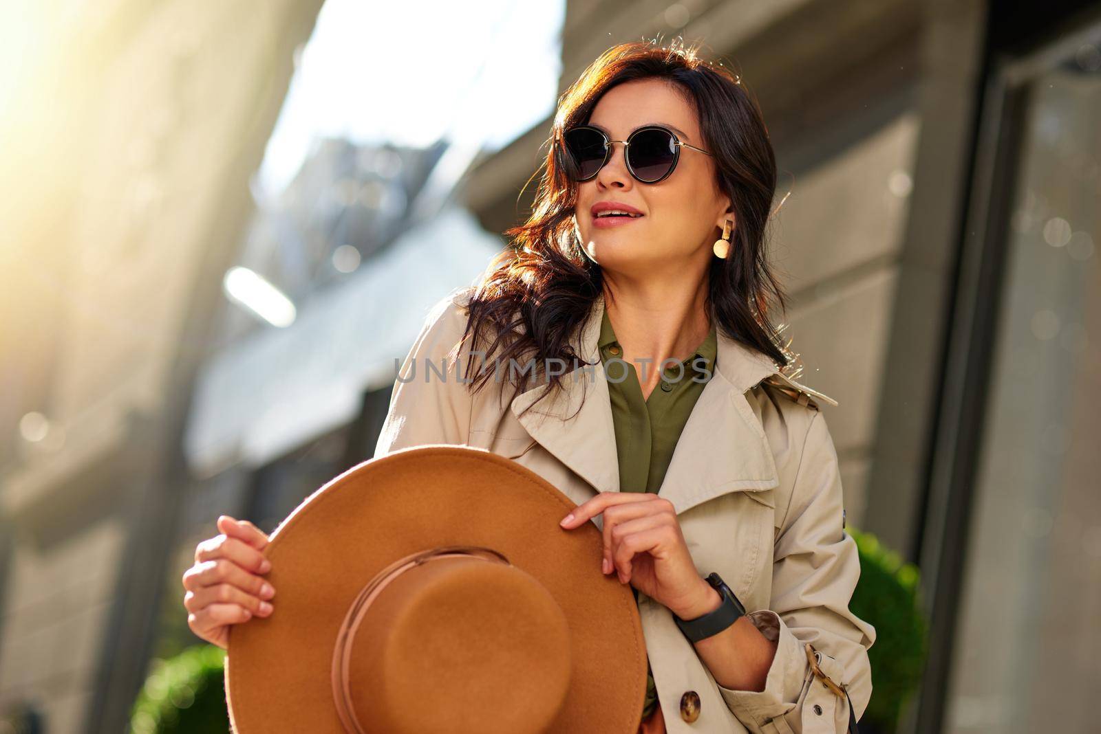 Young stylish woman wearing eyeglasses holding hat and looking aside while walking city streets on a warm autumn day by friendsstock