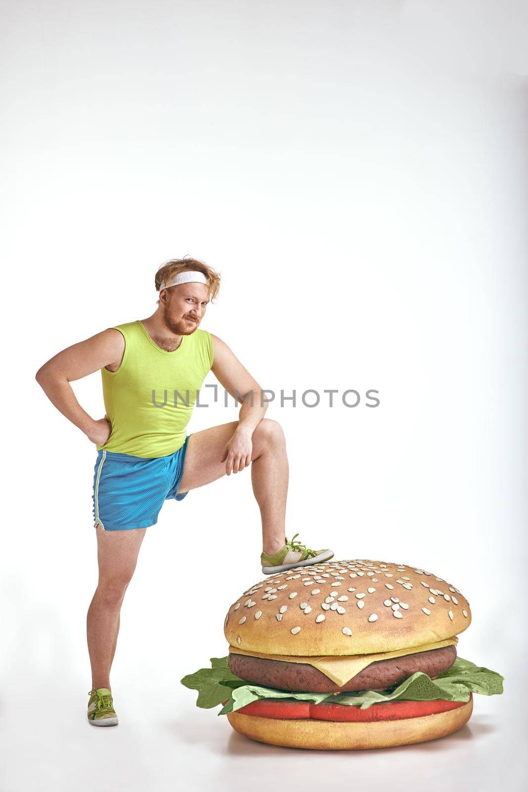 Funny picture of red haired, bearded, plump man on white background. Man wearing sportswear. Man put his leg to a huge sandwich