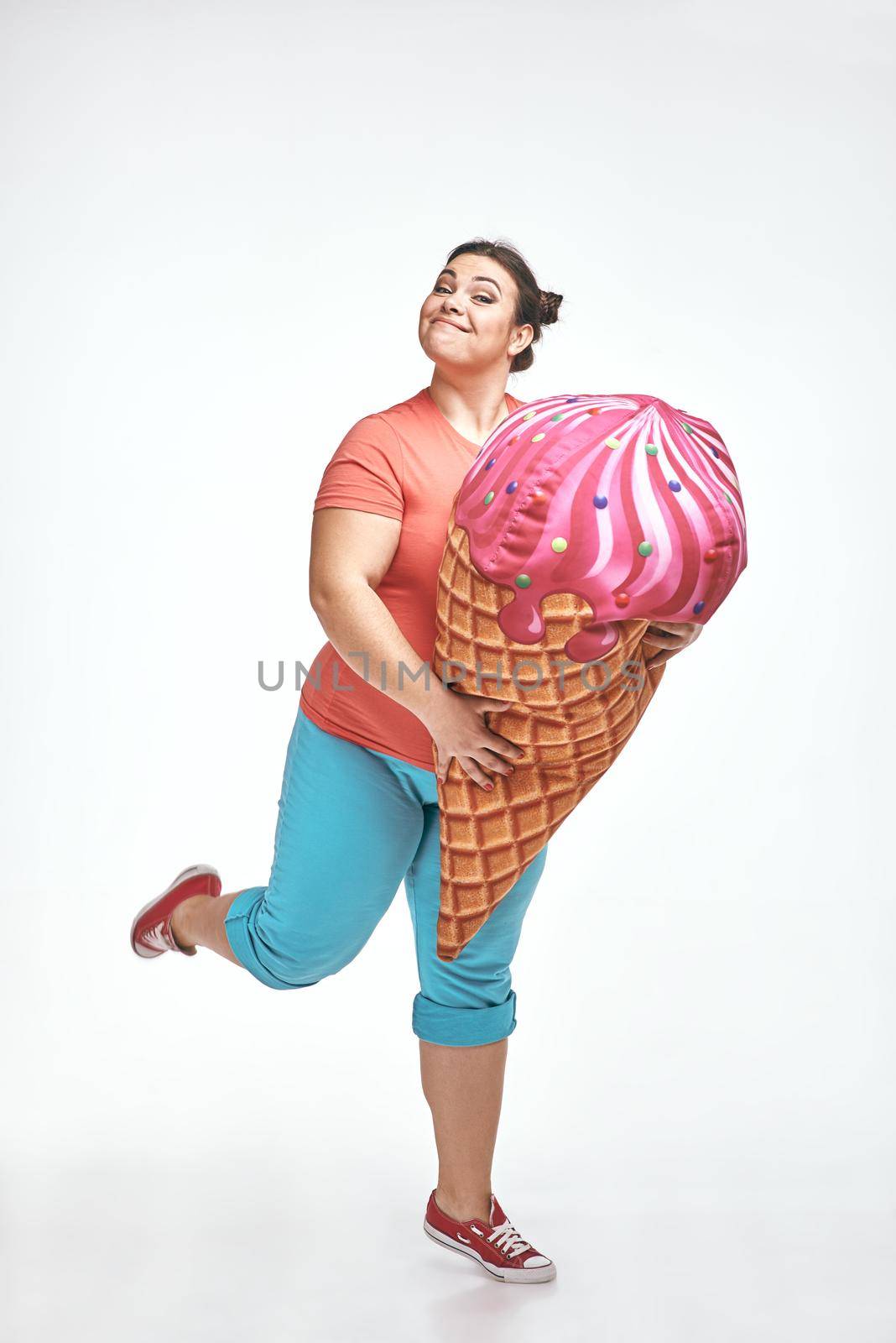 Funny picture of amusing, brunette, chubby woman on white background. Woman is holding a huge ice cream