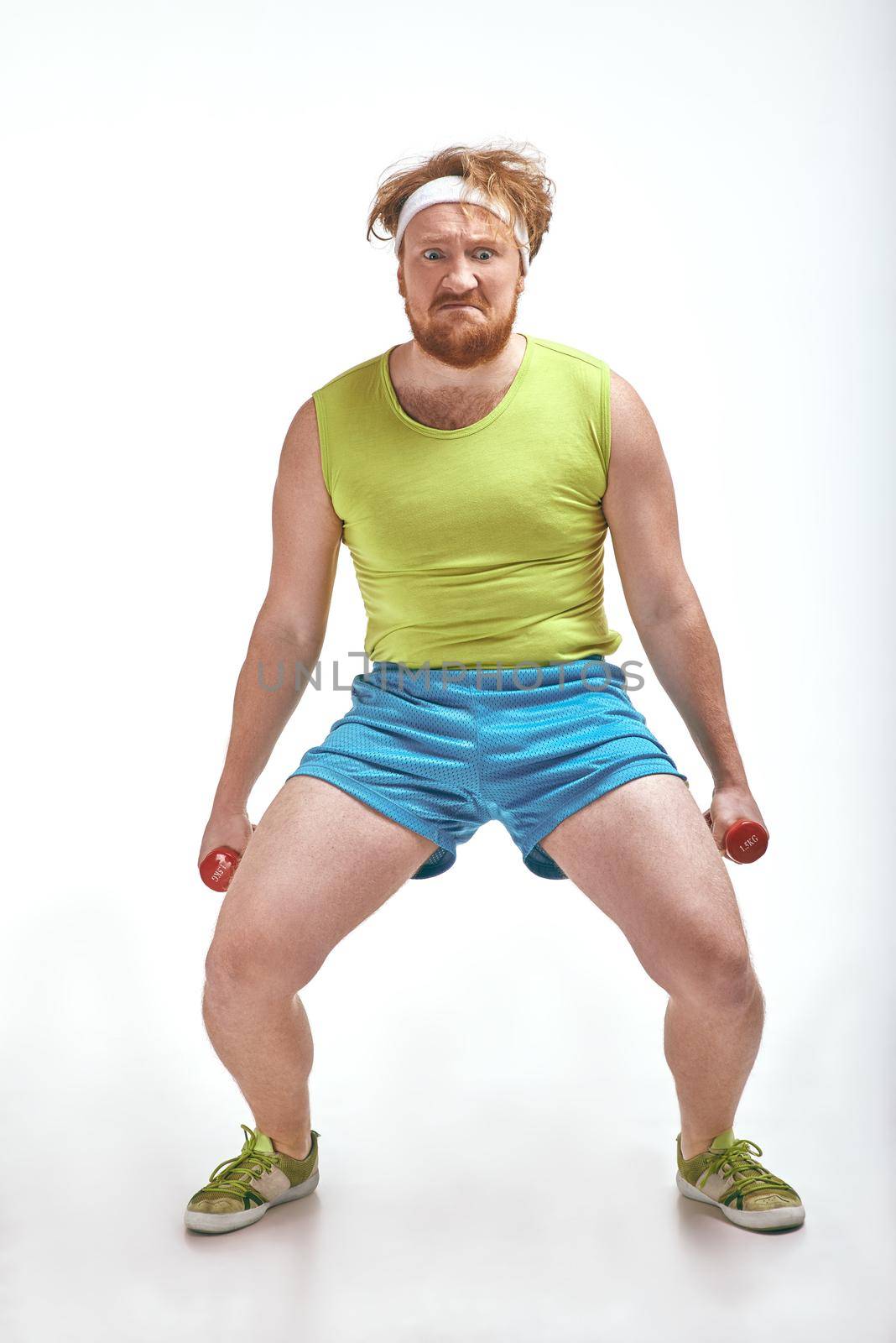 Funny picture of red haired, bearded, plump man on white background. Man wearing sportswear. Man holding the dumbbells