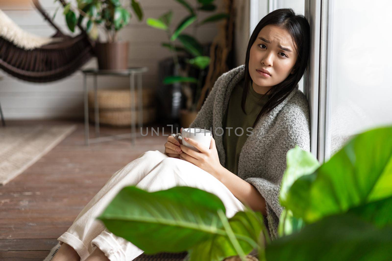 Sad sick asian girl sitting with coffee mug near window on floor, looking upset and unhappy.