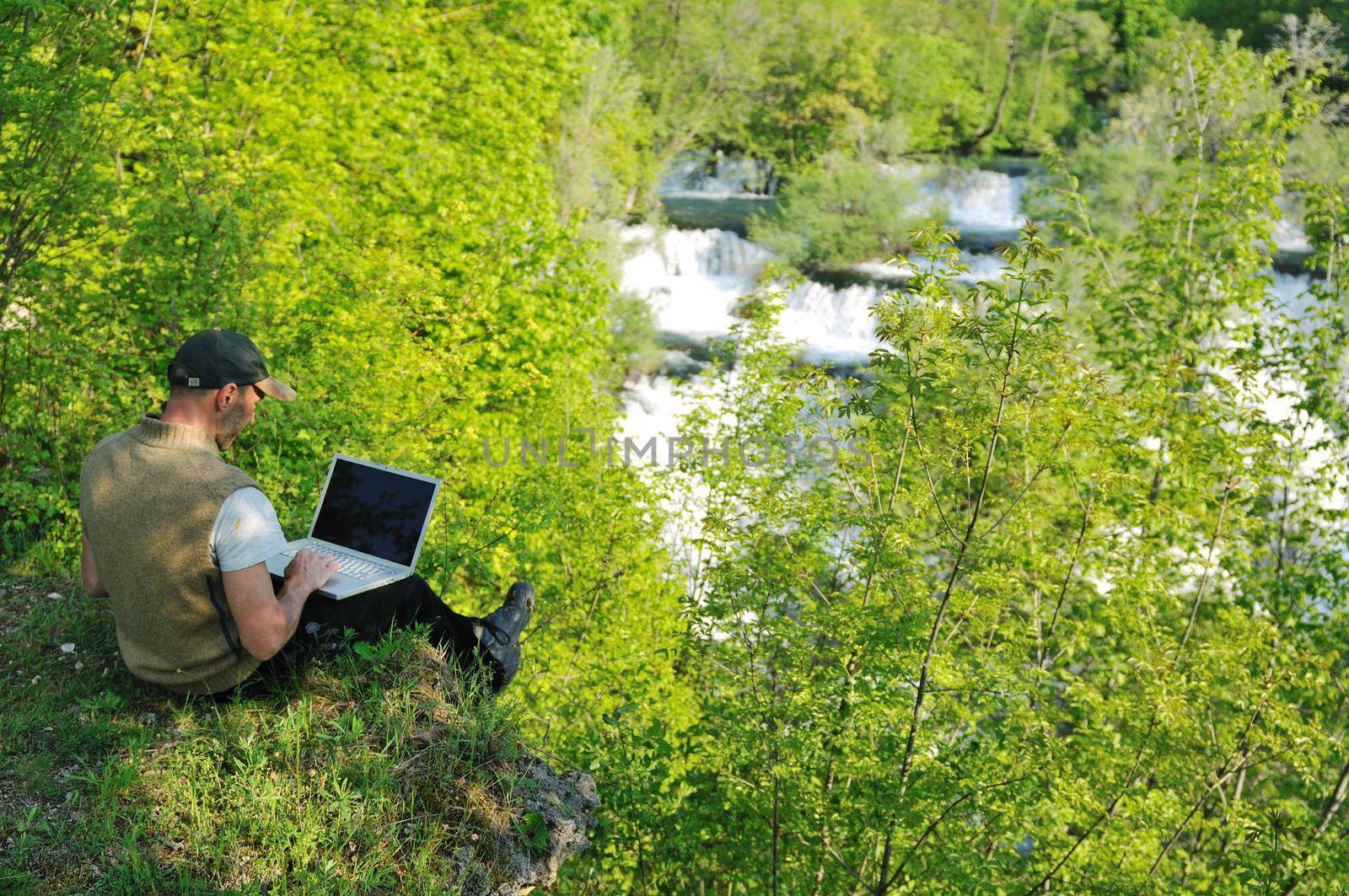 young businessman with hat work on thin laptop outdoor in nature with beautiful waterfalls in background representing freedom and wireless technology concept 