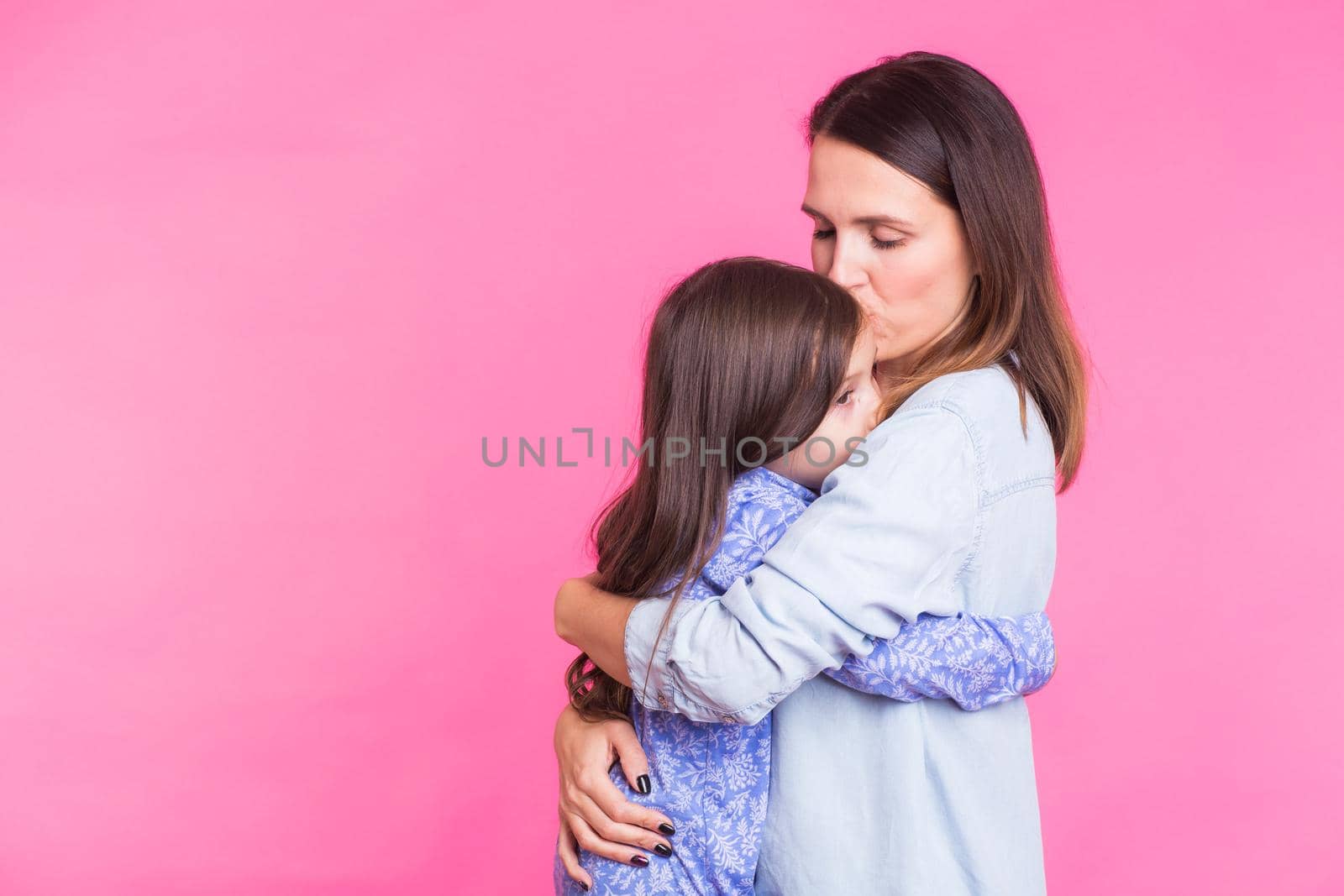 people, happiness, love, family and motherhood concept - happy little daughter hugging and kissing her mother over pink background.