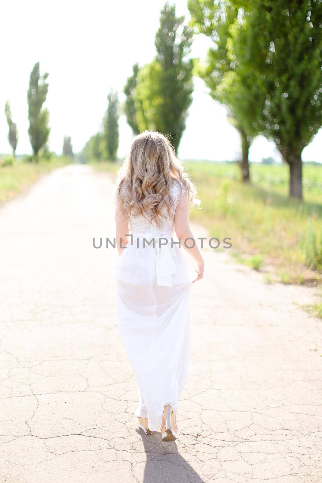 Back view of blonde bride wearing white dress walking on road. Concept of wedding photo session and fiancee.