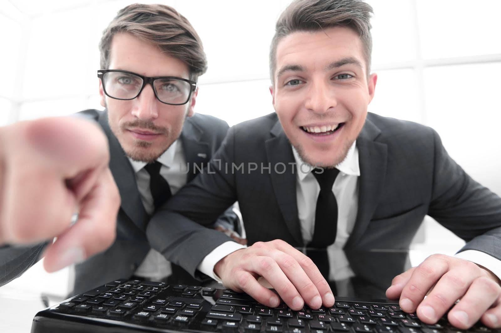 two young businessman are looking into the camera while sitting in the office. One shows the index finger