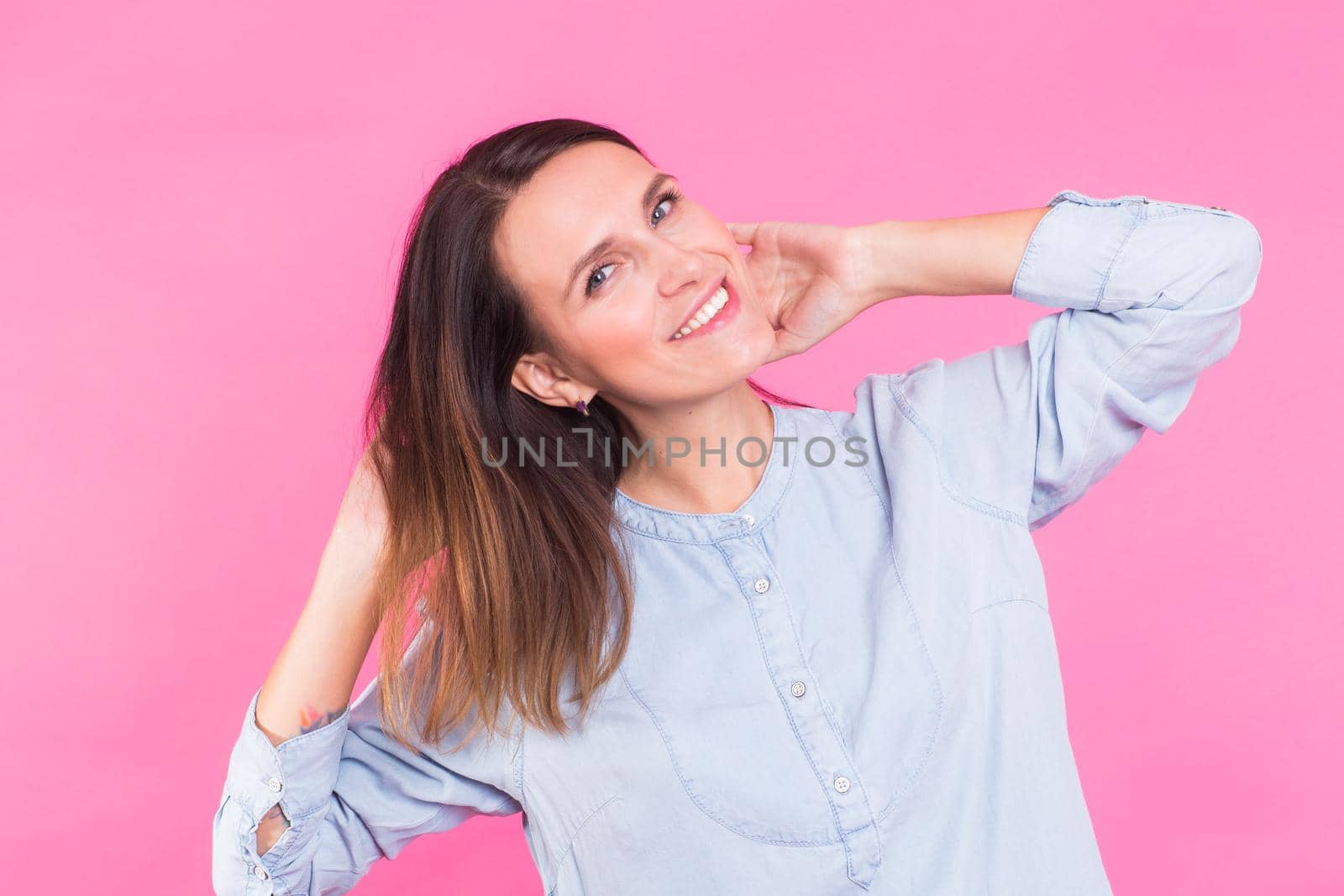 Portrait of a beautiful woman with long brown hair wearing blue cotton blouse, standing waist up smiling on a pink background.