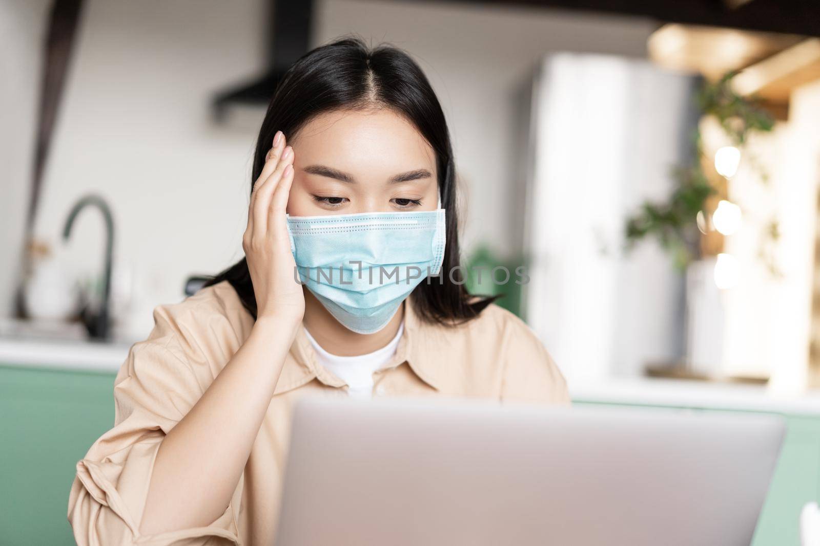 Young sick asian girl in medical face mask using laptop, working from home on quarantine by Benzoix