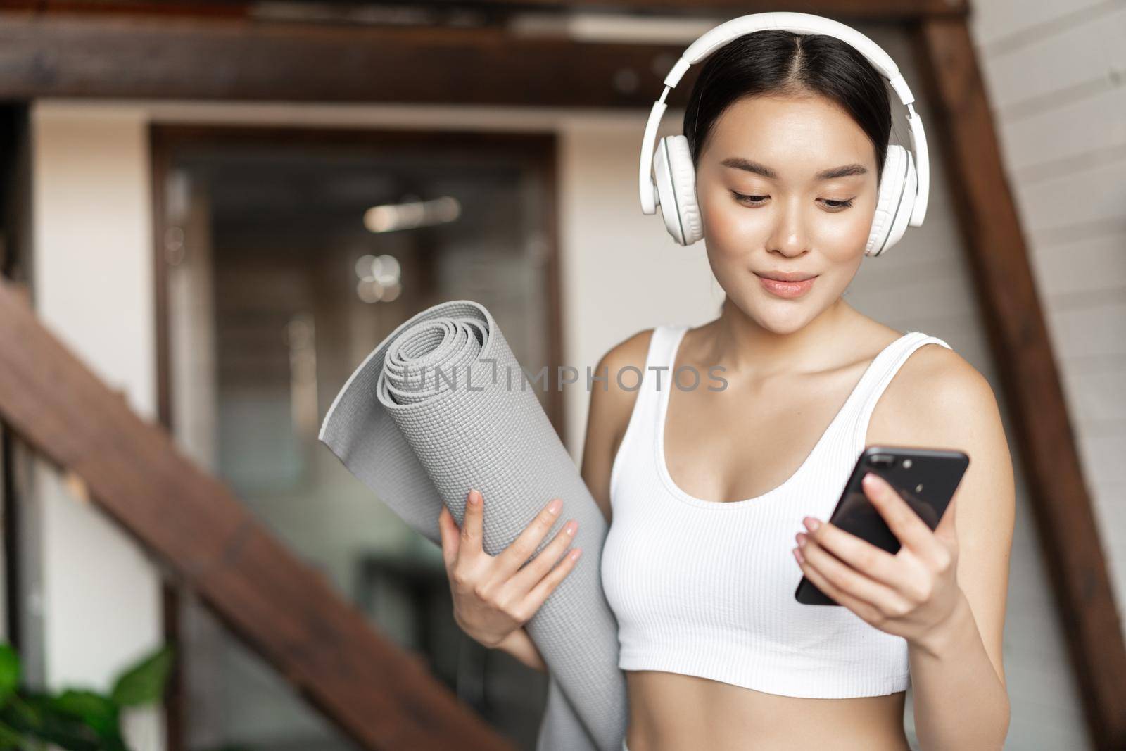 Young asian woman standing at home in headphones, listening music and going workout on floor mat, practice yoga or meditate in living room by Benzoix