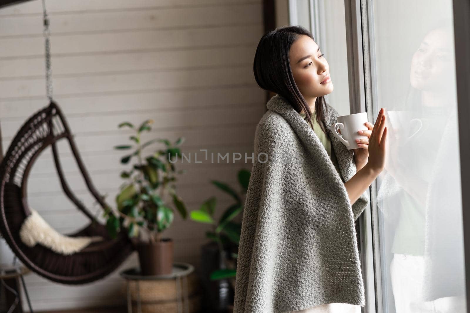 Romantic asian woman touching window, looking outside with thoughtful face, drinking tea and gazing nostalgic, staying at home.