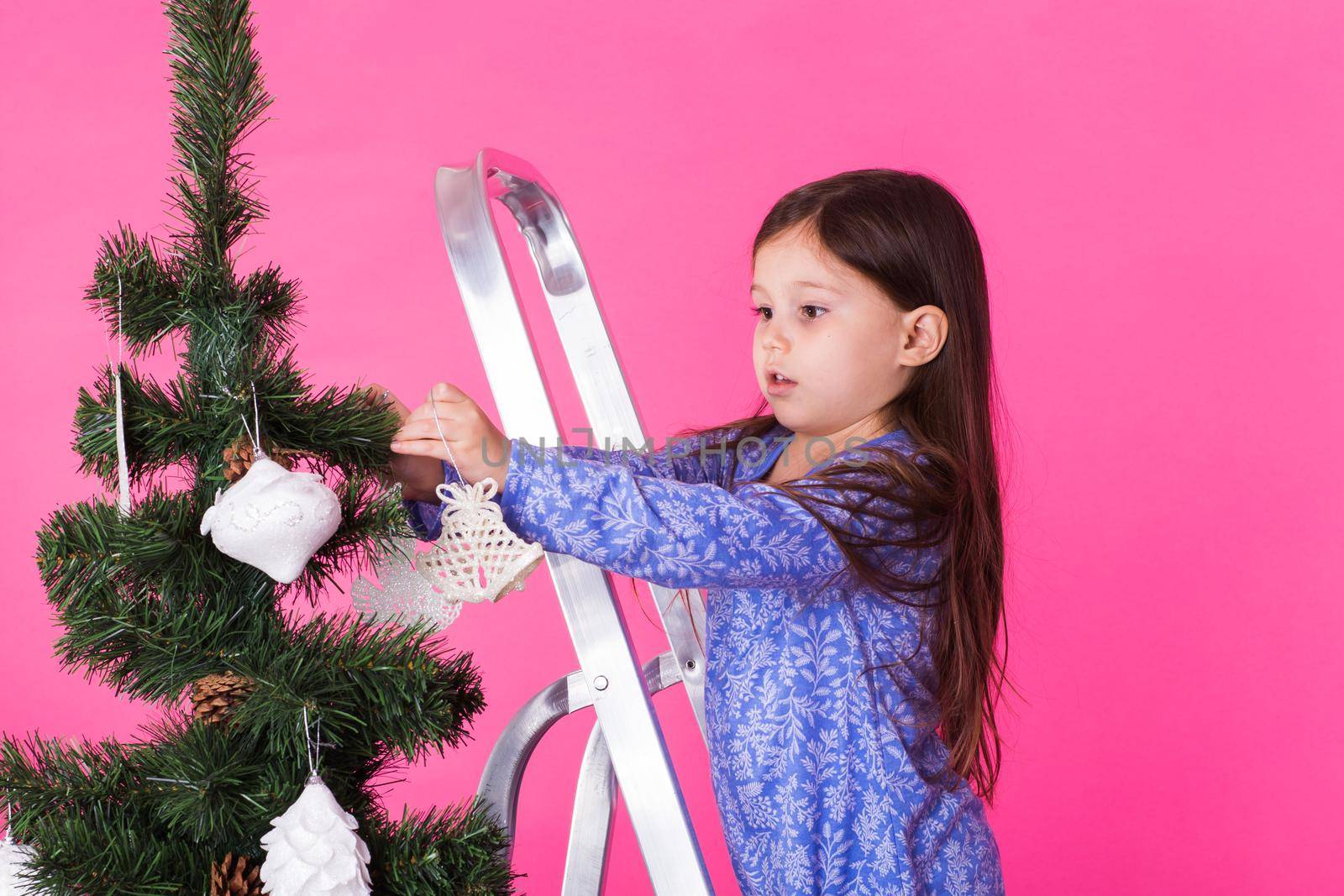 Children, holidays and christmas concept - little girl decorating christmas tree on pink background