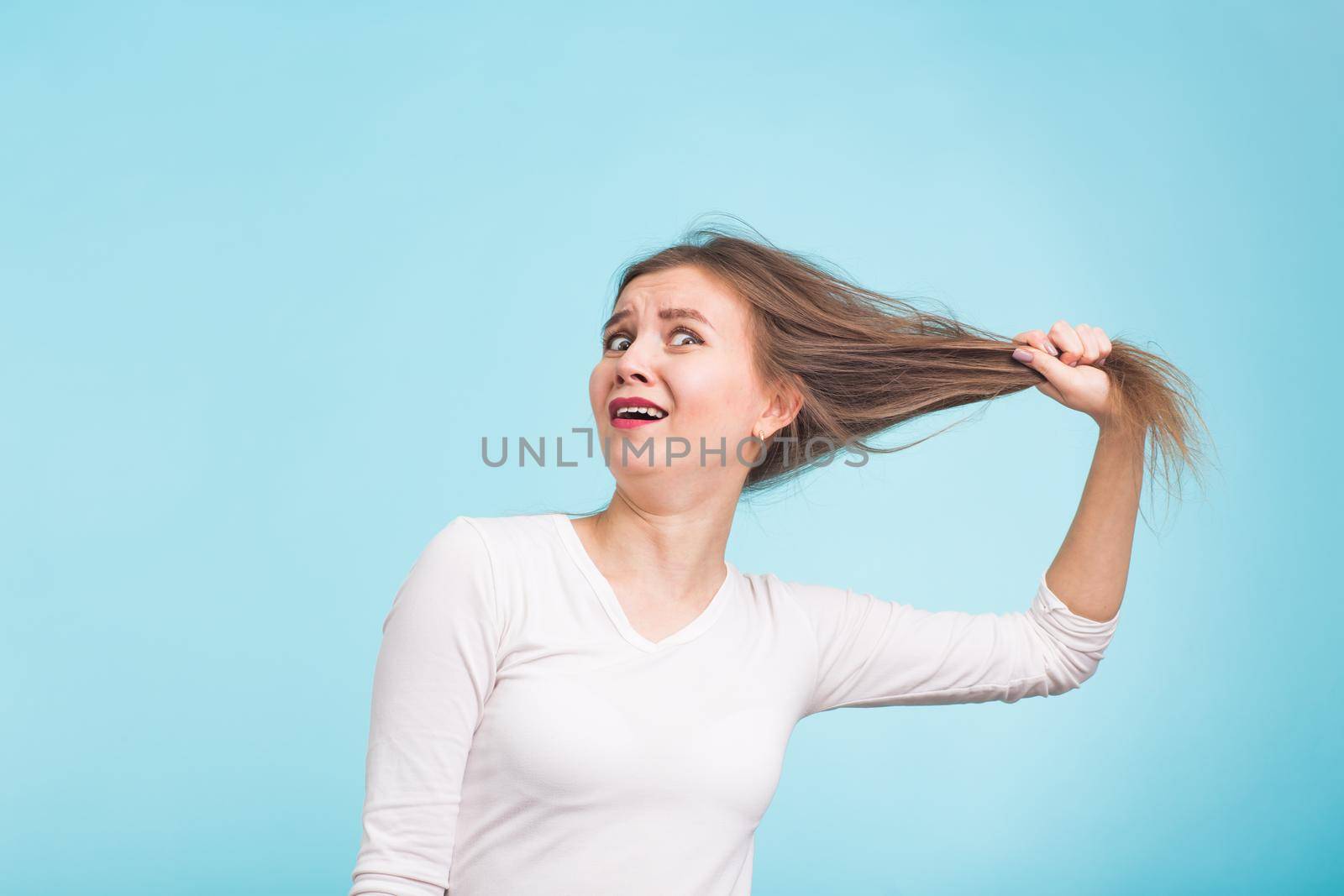 Scared young woman with unhealthy long hair keep hair in hand with nature make up portrait isolated on blue by Satura86