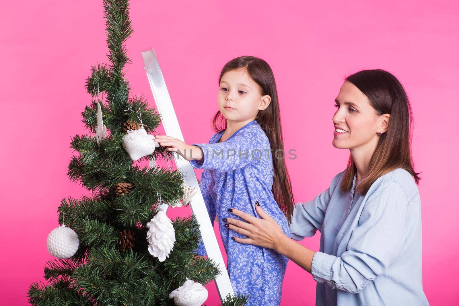 Holidays, family and christmas concept - mother and daughter decorating christmas tree on pink background