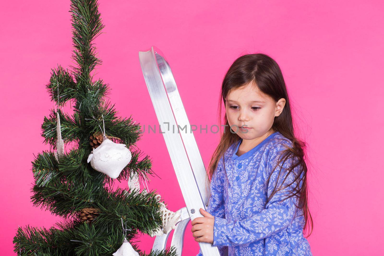 Children, holidays and christmas concept - little girl decorating christmas tree on pink background