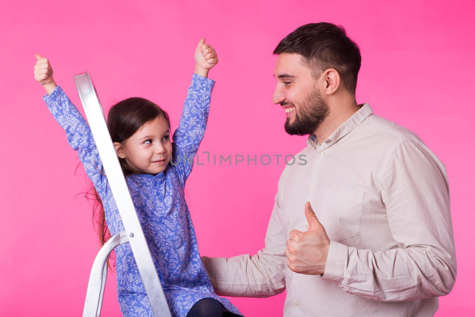 Father and her little daughter with thumbs up over pink background. Adult man and baby girl are happy.