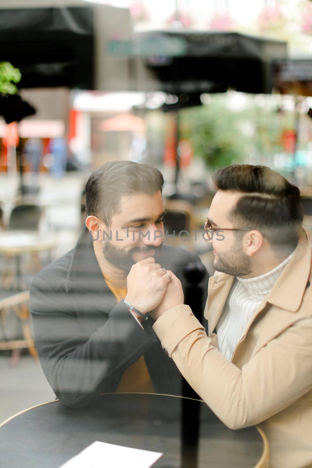 Reflection in glass of two young handsome gays talking at street cafe and holding hands. Concept of same sex couple and lgbt.
