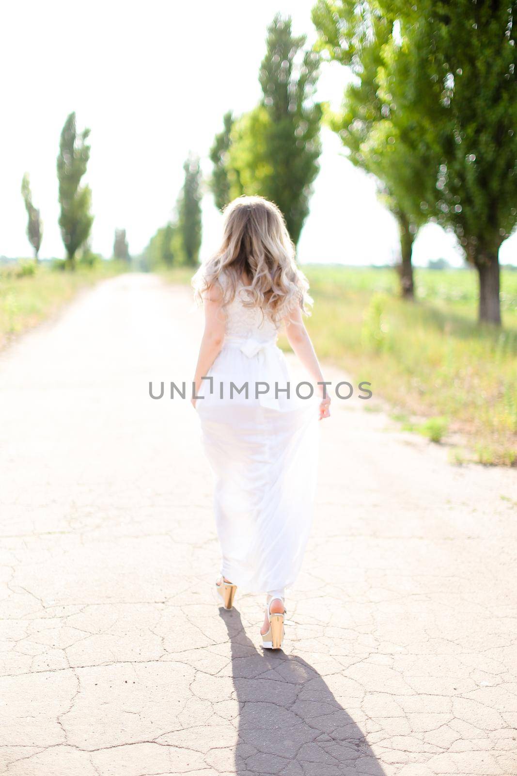 Back view of blonde caucasian bride wearing white dress walking on road. Concept of wedding photo session and fiancee.