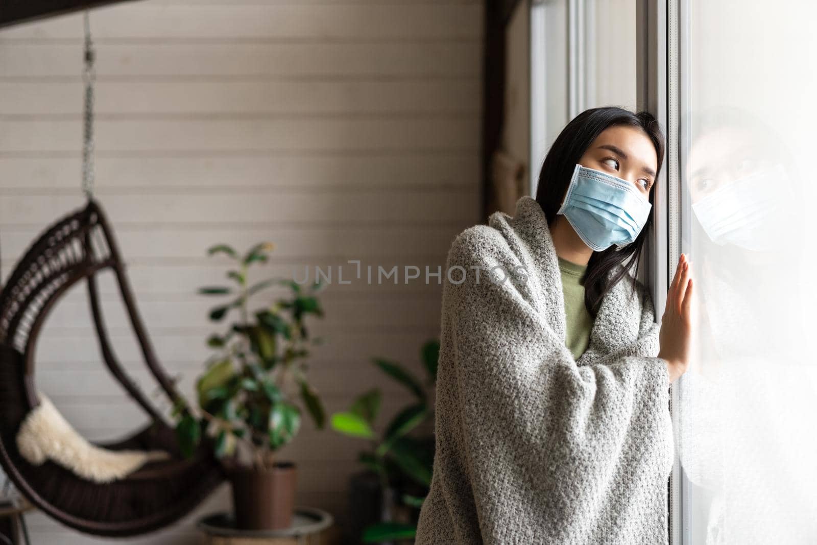 Sad asian girl in medical face mask looking outside window and yearning go for walk, being ill, catching covid 19 or flu by Benzoix