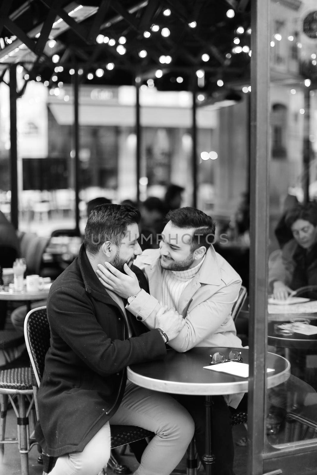 Black and white photo of caucasian gays sitting at street cafe and hugging. Concept of relationship and same sex couple.