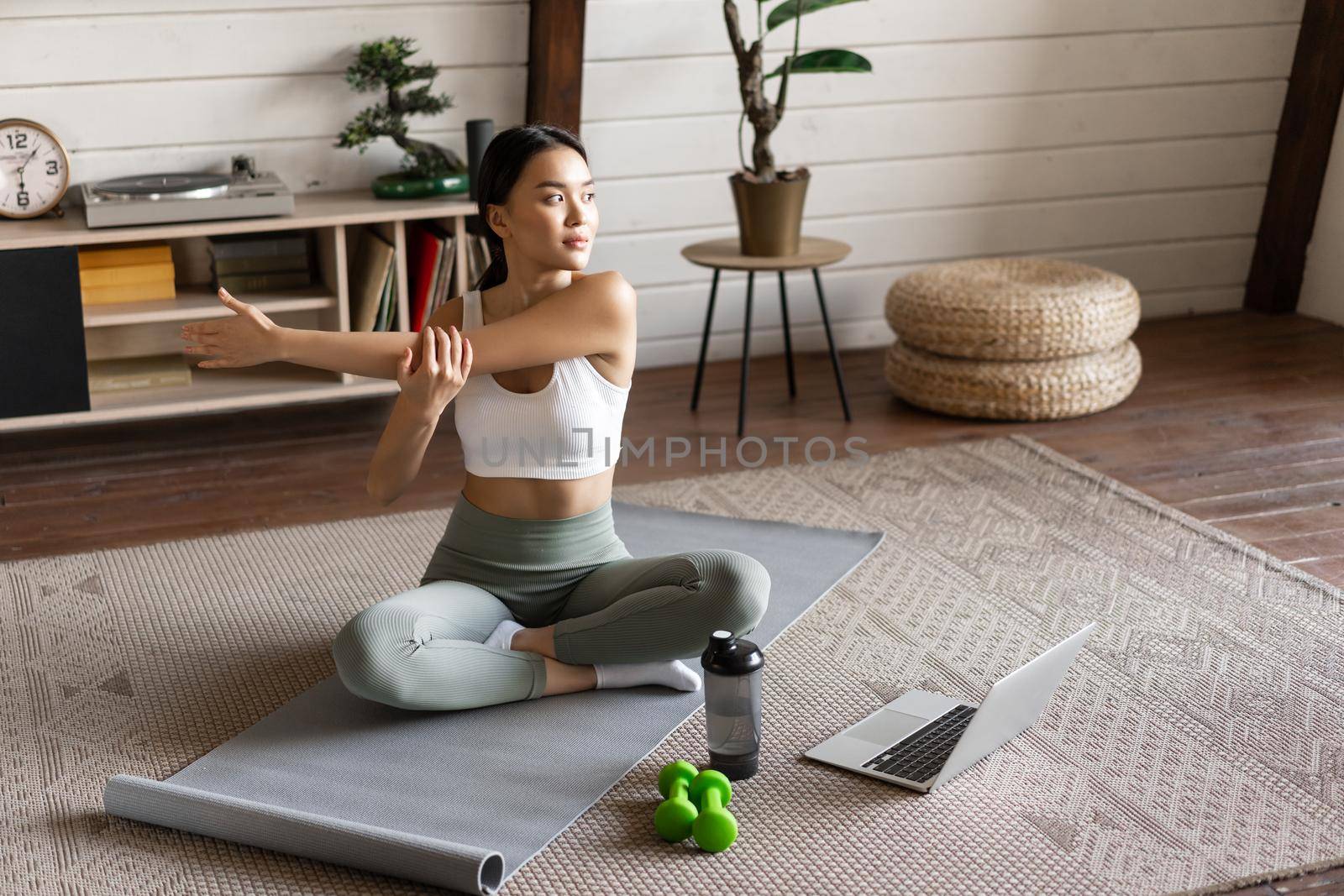Young asian fitness girl doing stretching, sport workout at home, stretch arms and looking at laptop, follow exercise video on computer.